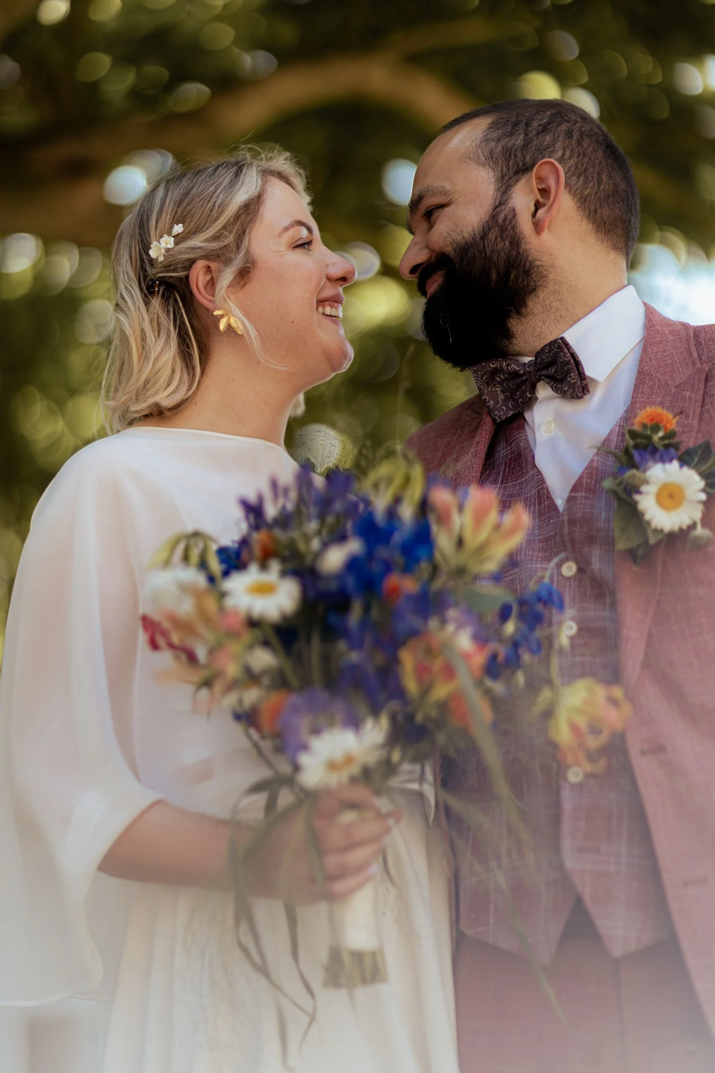 Een getrouwd paar kijkt elkaar lachend aan tijdens hun bruiloft, omringd door groene bomen.