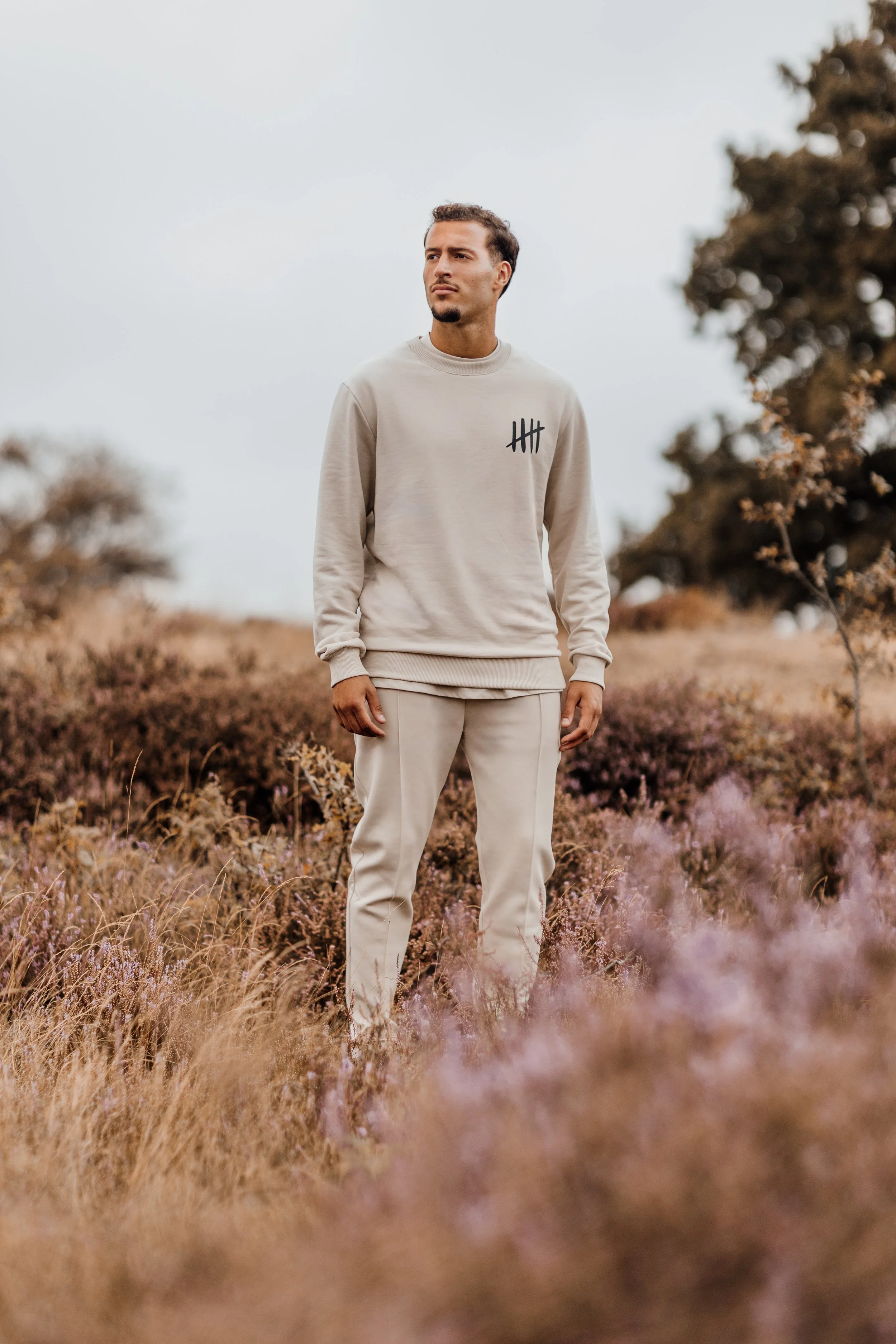 Man in beige joggingpak terwijl hij in een veld met droge, paarse en bruine planten staat, onder een lichte grijze lucht.