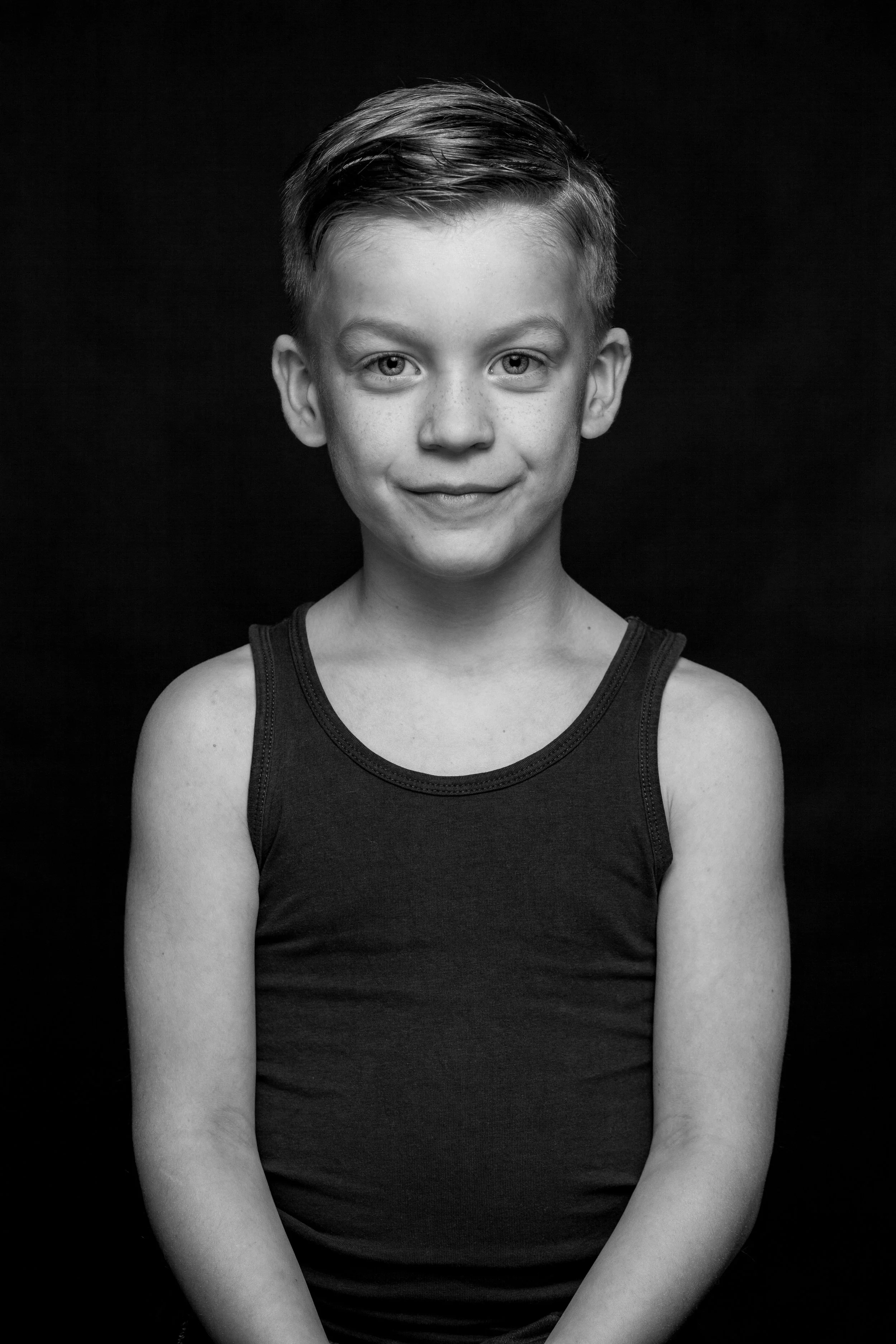 Zwarte en witte foto van een jongen met een kort getrimd haar, glimlach, in een zwart tanktop, tegen een zwarte achtergrond.