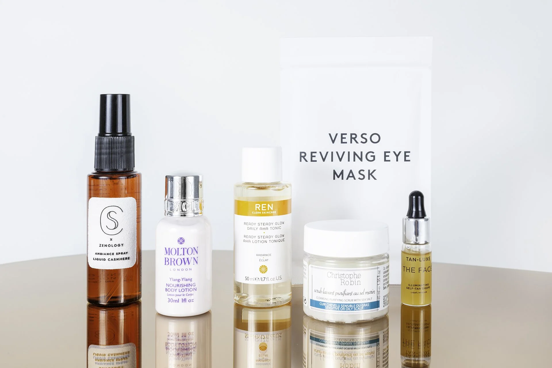Gezichtsverzorgingsproducten en een gezichtsmasker op een glazen oppervlak, inclusief een spray, body lotion, toner, scrub, serum en een oogmasker, met een VERSO REVIVING EYE MASK-verpakking op de achtergrond.