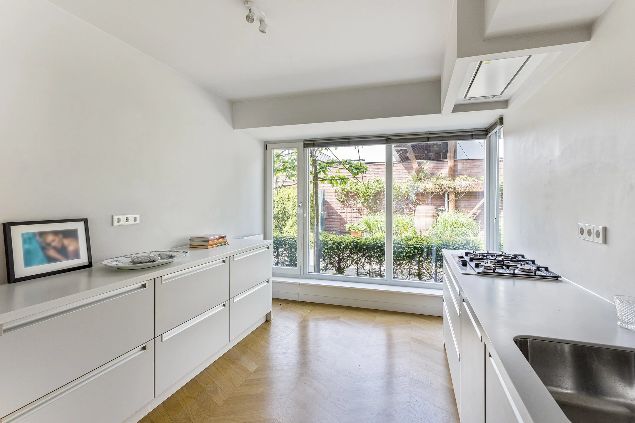 Een moderne witte keuken met grote glazen ramen die uitkijken op een tuin met groen begroeiing.