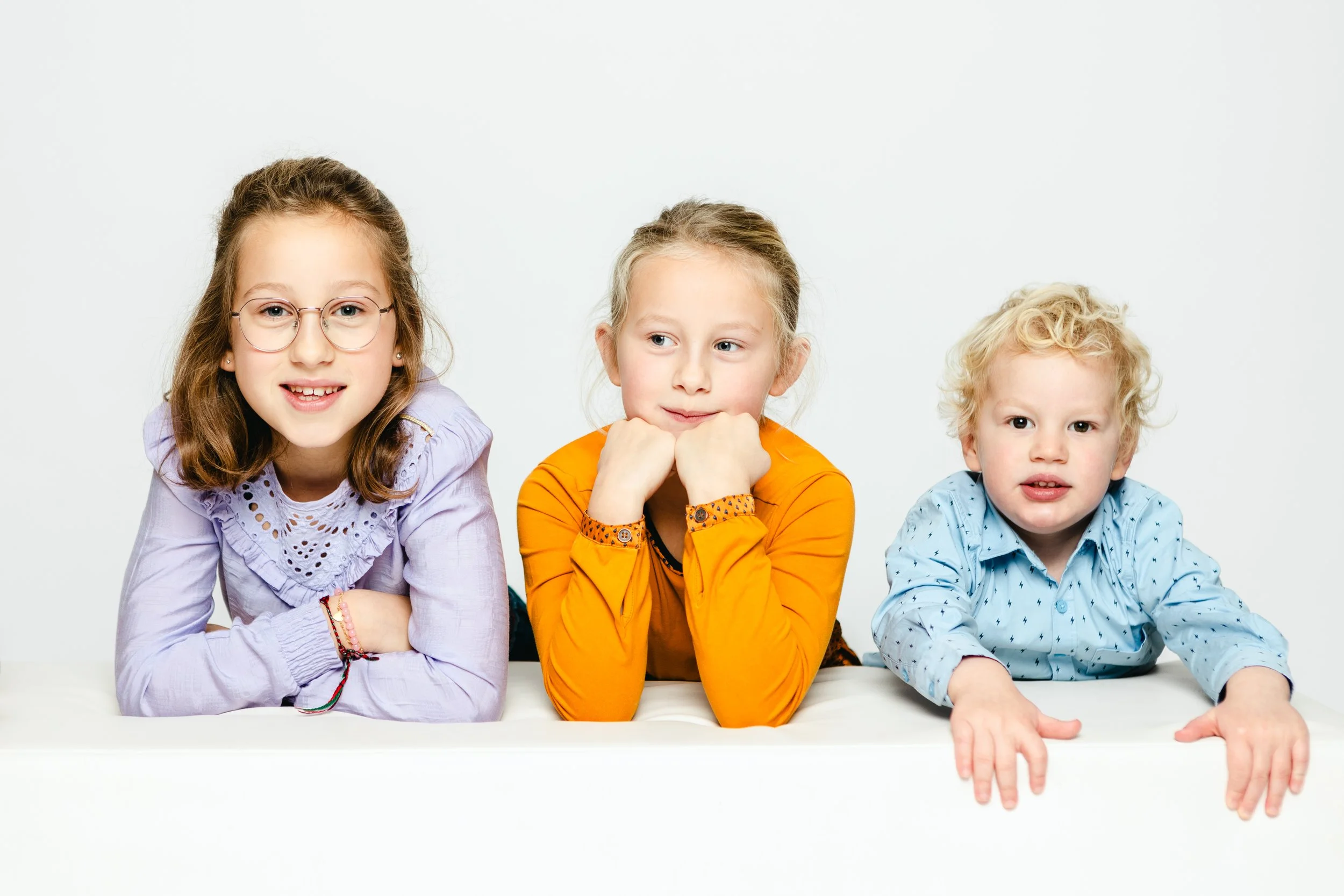 Drie kinderen liggen op een witte tafel tegen een witte achtergrond, lachend en kijken naar de camera.