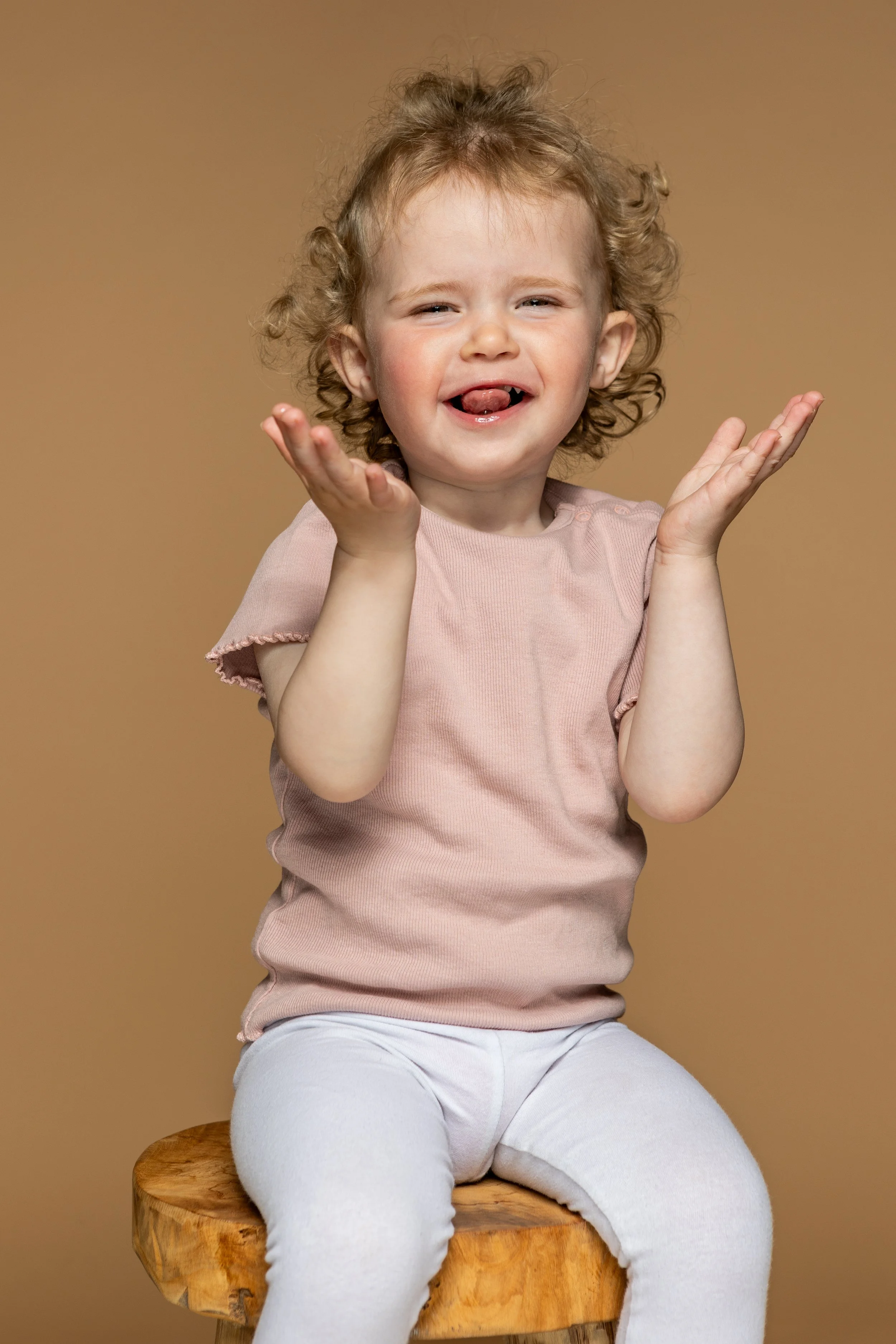 Lachende jong meisje met gekrulde haar, biddend met handgebaren, zittend op een houten kruk, tegen een beige achtergrond.