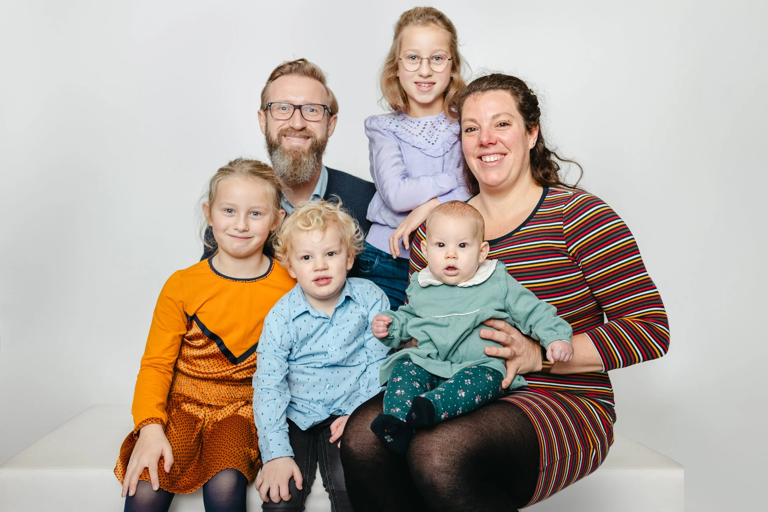 Gezicht van een gezin met vier kinderen en twee volwassenen, zittend op een wit platform voor een witte achtergrond.