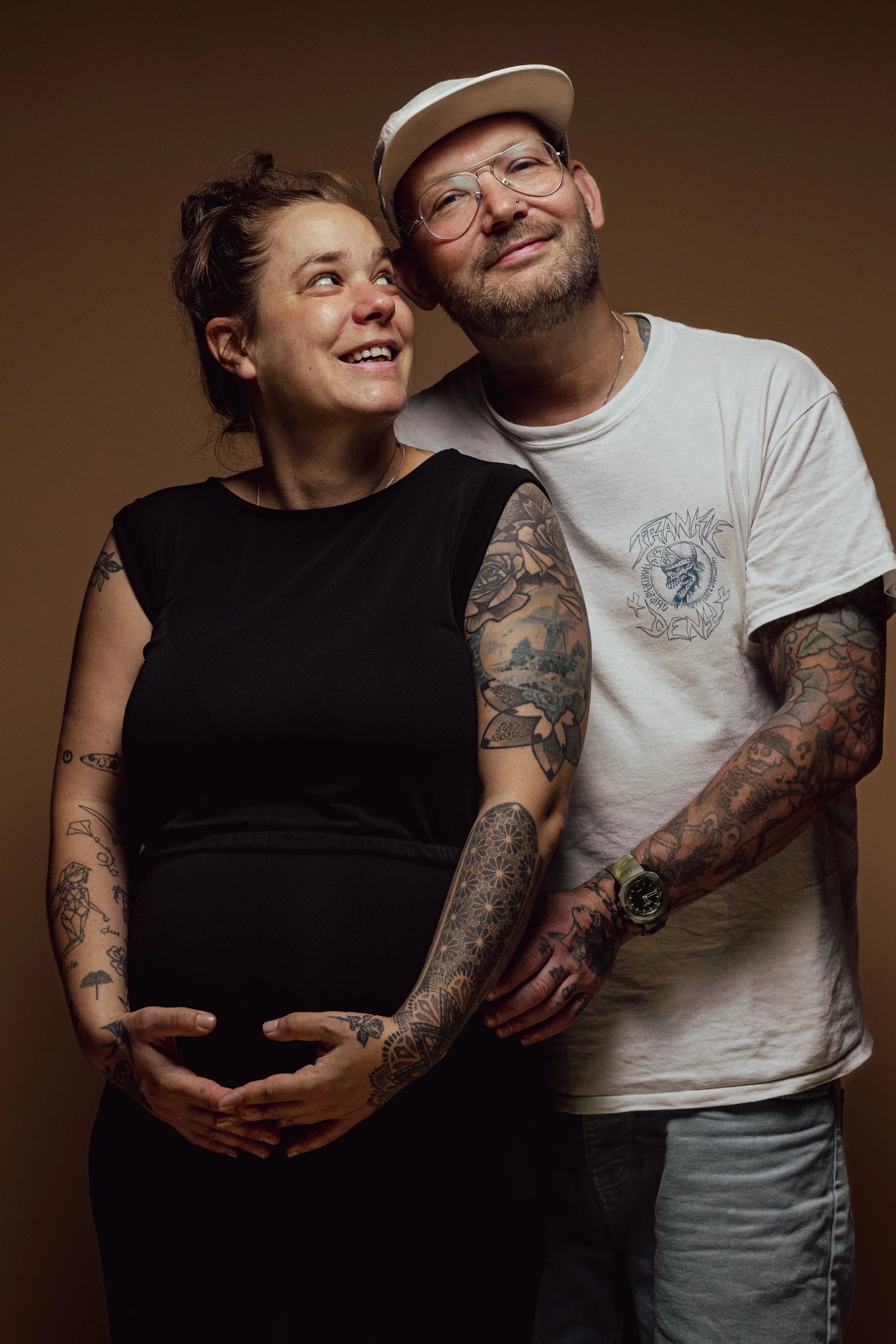 Een zwangere vrouw met tattoos die glimlacht en een man met tattoos die naast haar staat, beiden poseren tegen een effen achtergrond.