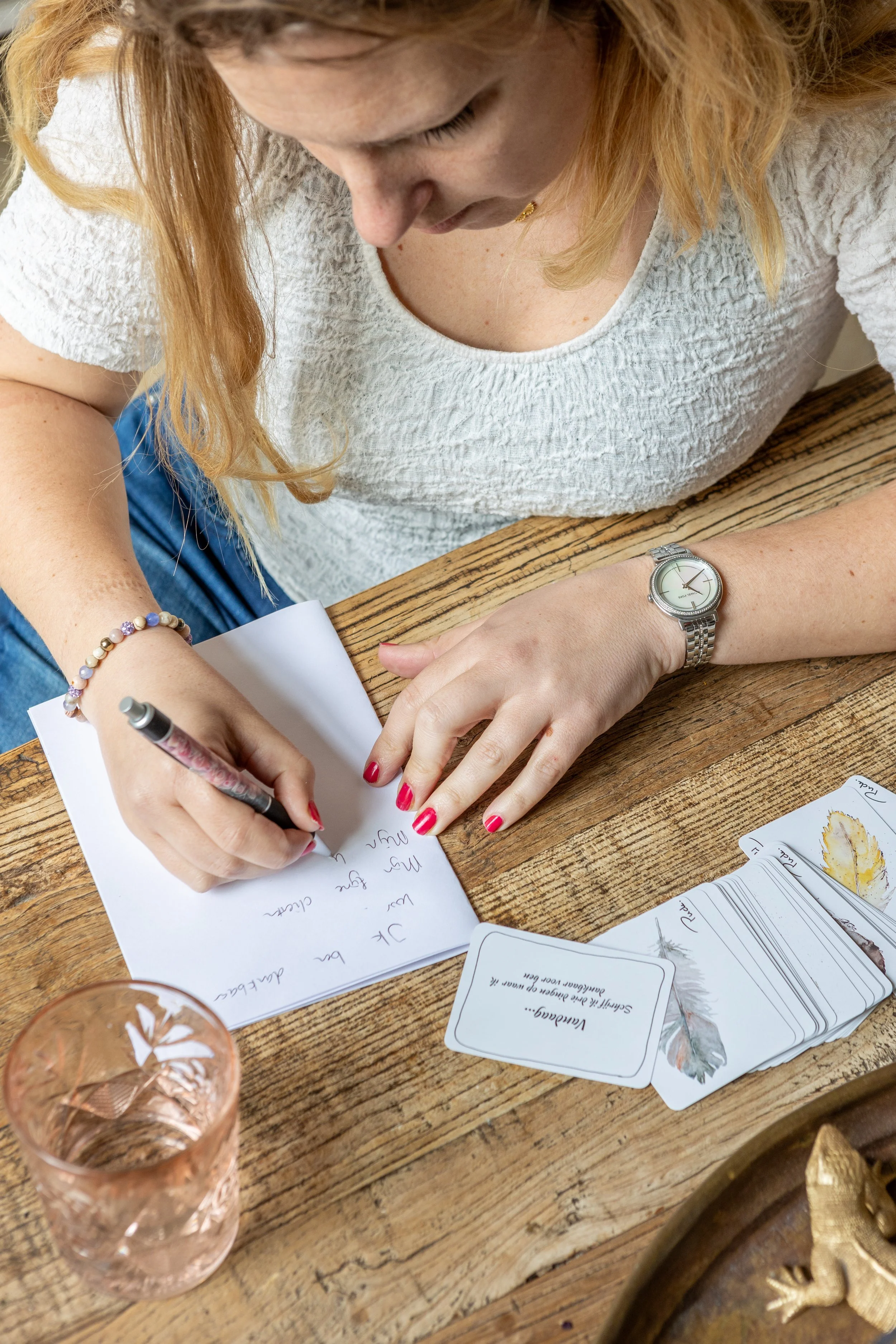 Vrouw schrijft in een notitieboekje op een houten tafel, met kaarten en een glas water voor haar.