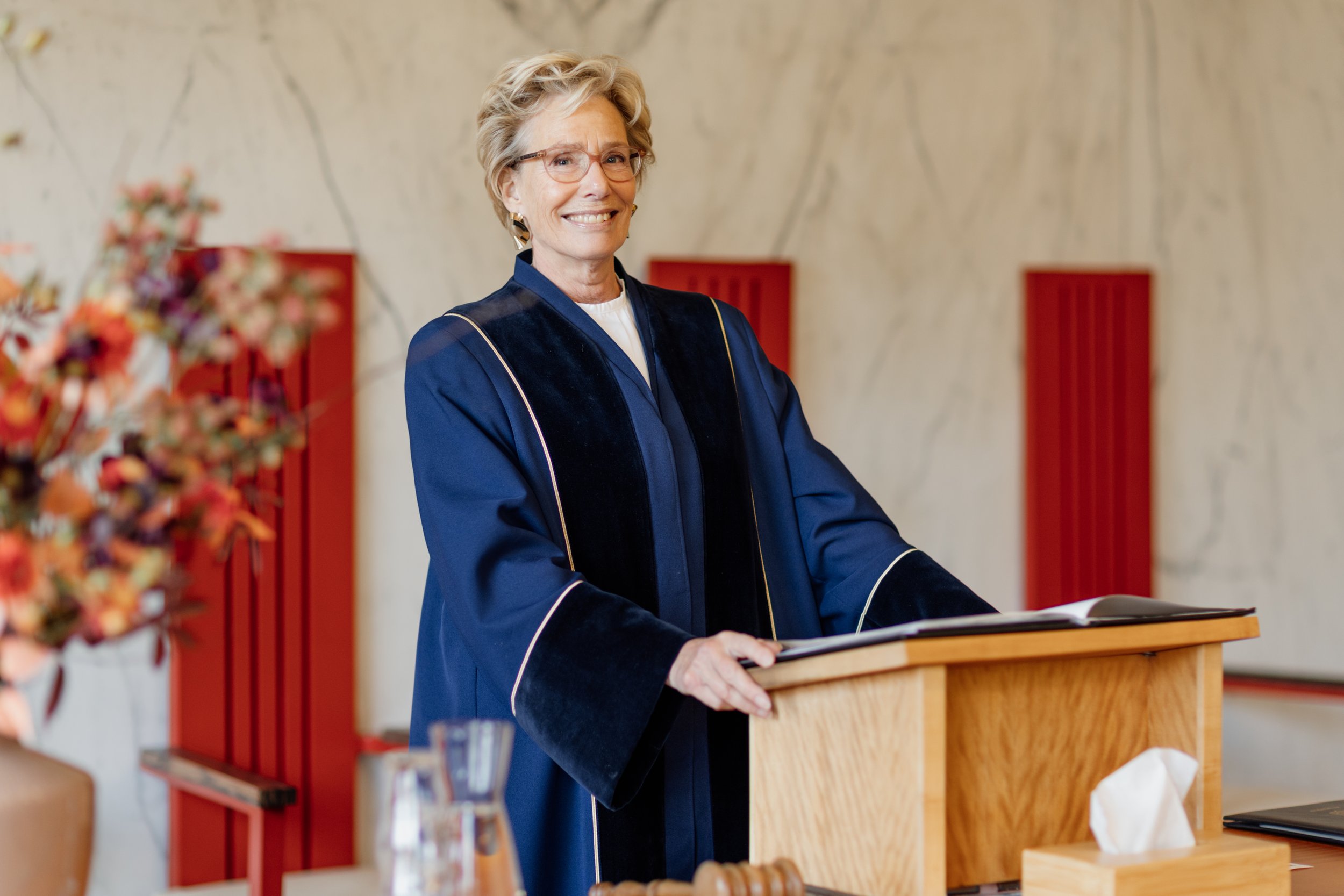 Een vrouw in een universitaire toga staat achter een lessenaar en glimlacht.