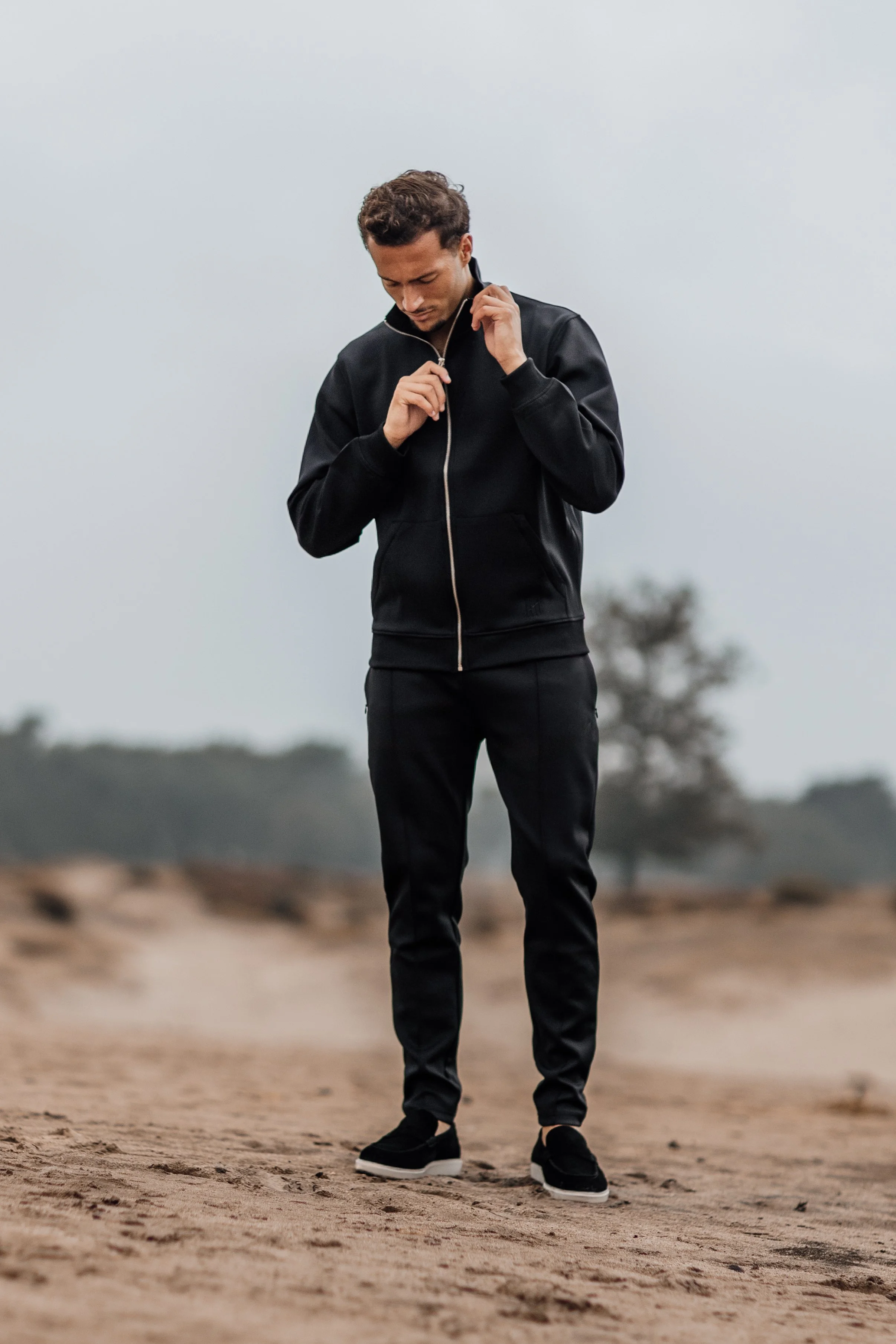 Man in zwart jasje en broek, loopt op aarde-achtig terrein, kijkt naar beneden.