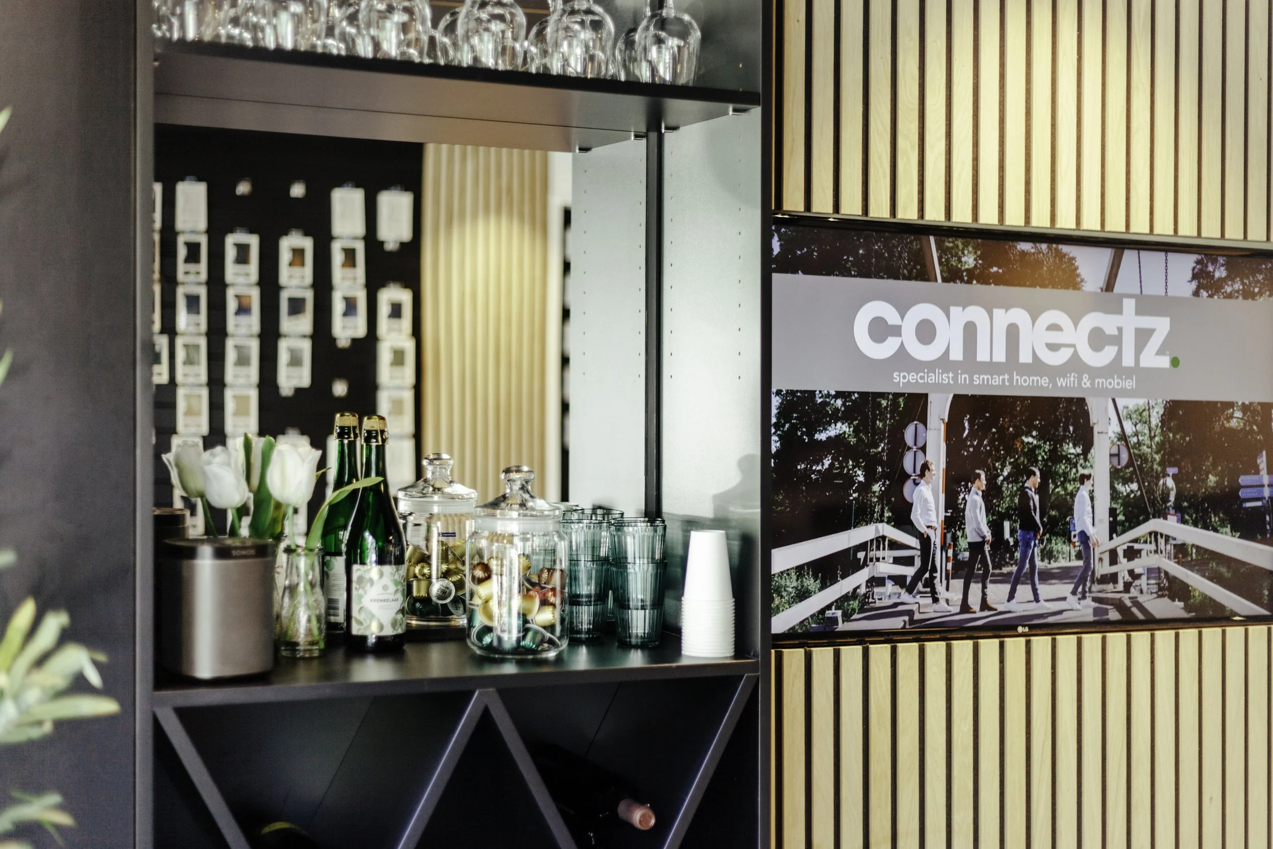 Binnenkant van een koffiebar met glazen, sapflessen, en bloemen op een zwart rek, naast een scherm met de tekst "connectz. specialist in smart home, wifi & mobiel".