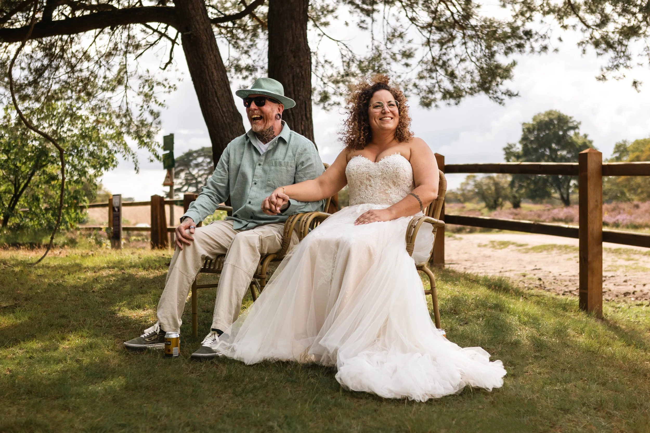 Een lachend bruid en bruidegom zitten samen op een bankje in een park, omgeven door bomen en tuin. Ze houden handen vasthouden en lijken te genieten van een gelukkige dag.