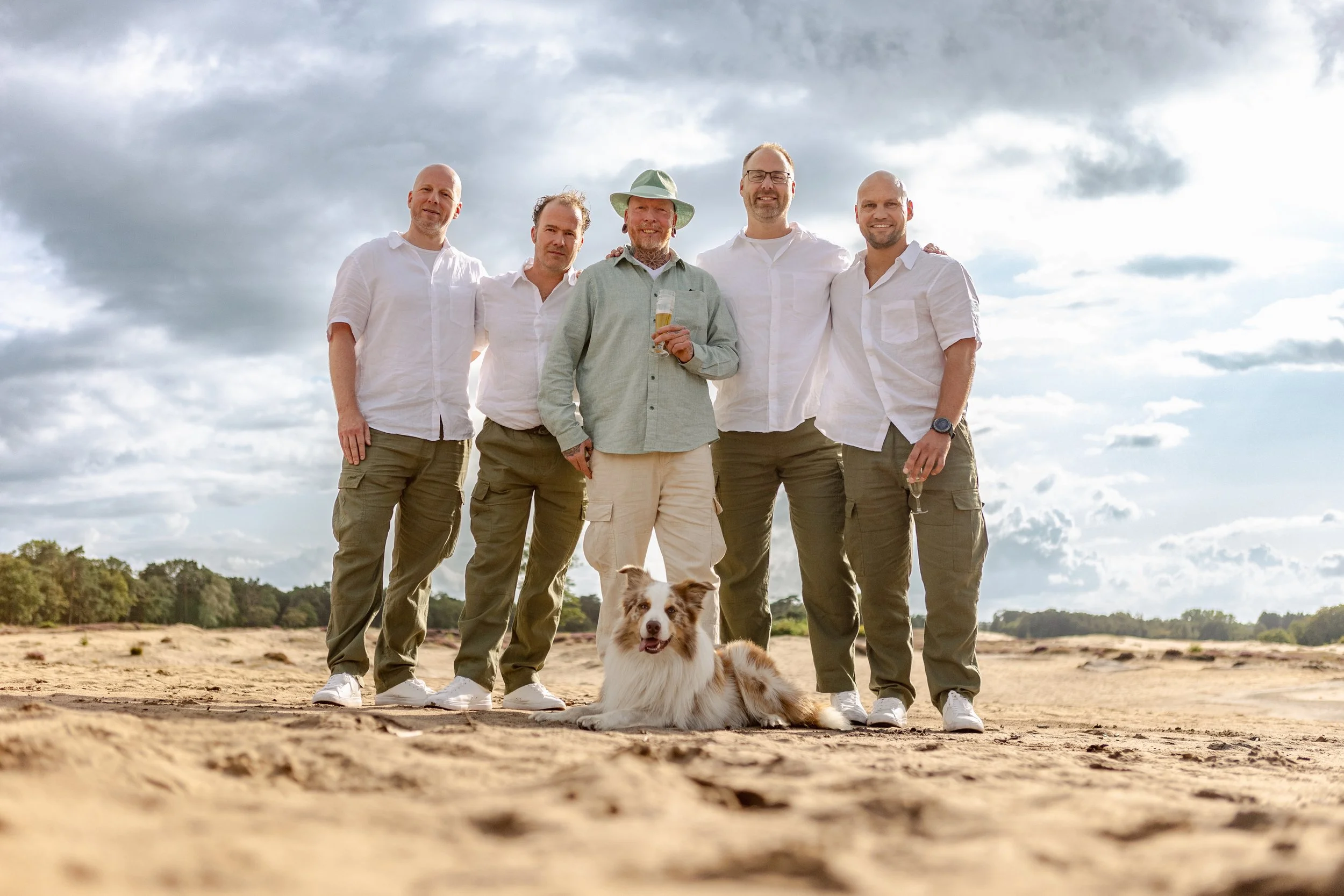 Vijf mannen en een Border Collie hond op een open veld, onder een bewolkte hemel, met een man die een glas champagne vasthoudt, allemaal gekleed in witte en aardekleurige kleding.