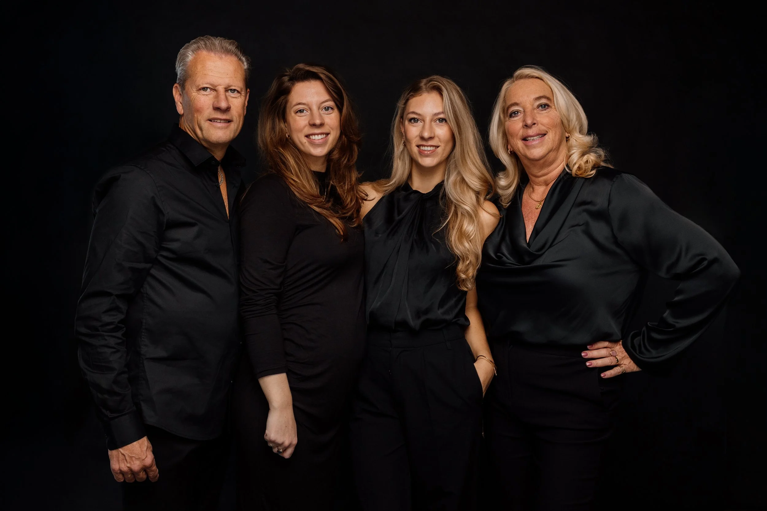 Vijf mensen standing samen tegen een zwarte achtergrond, gekleed in zwarte kleding, glimlachend.