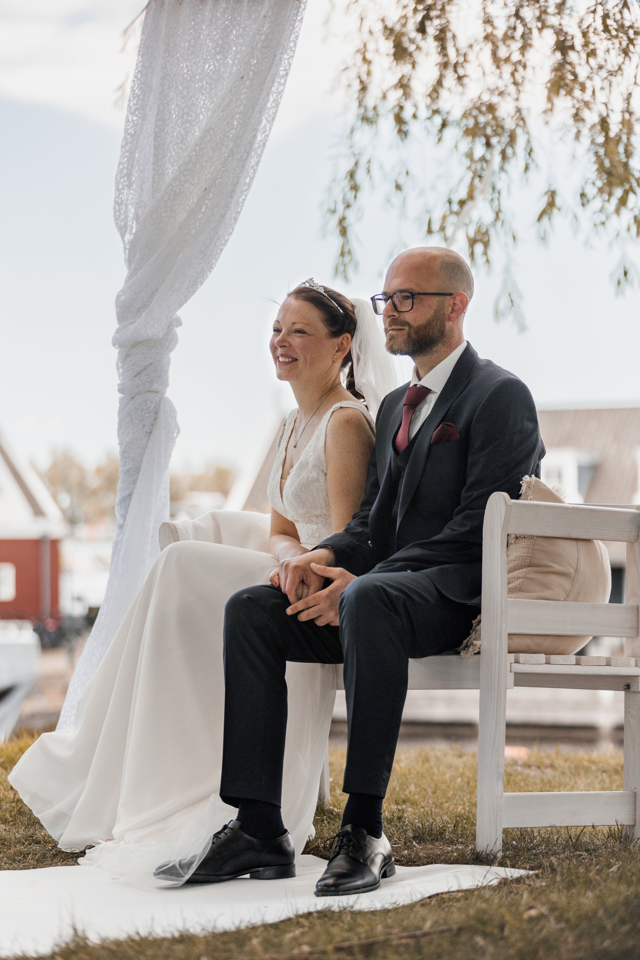 Een bruidskoppel zit samen tijdens hun huwelijk op een bankje onder een witte luifel buiten.