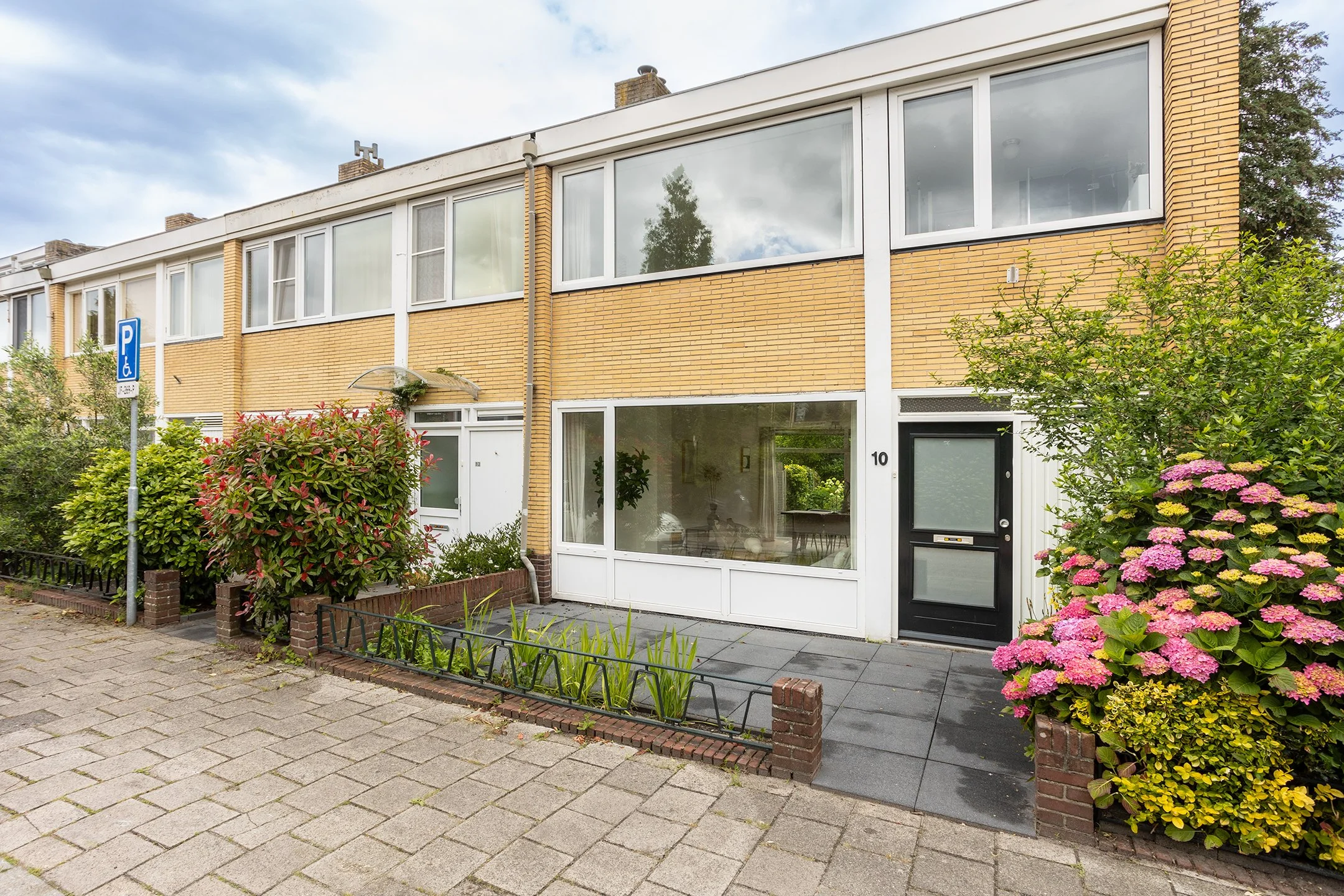 Gevel van een rijtjeshuis met grote ramen en tuin met bloemen en struiken, parkeerplek met parkeerschijf, en een parkeersymbool.