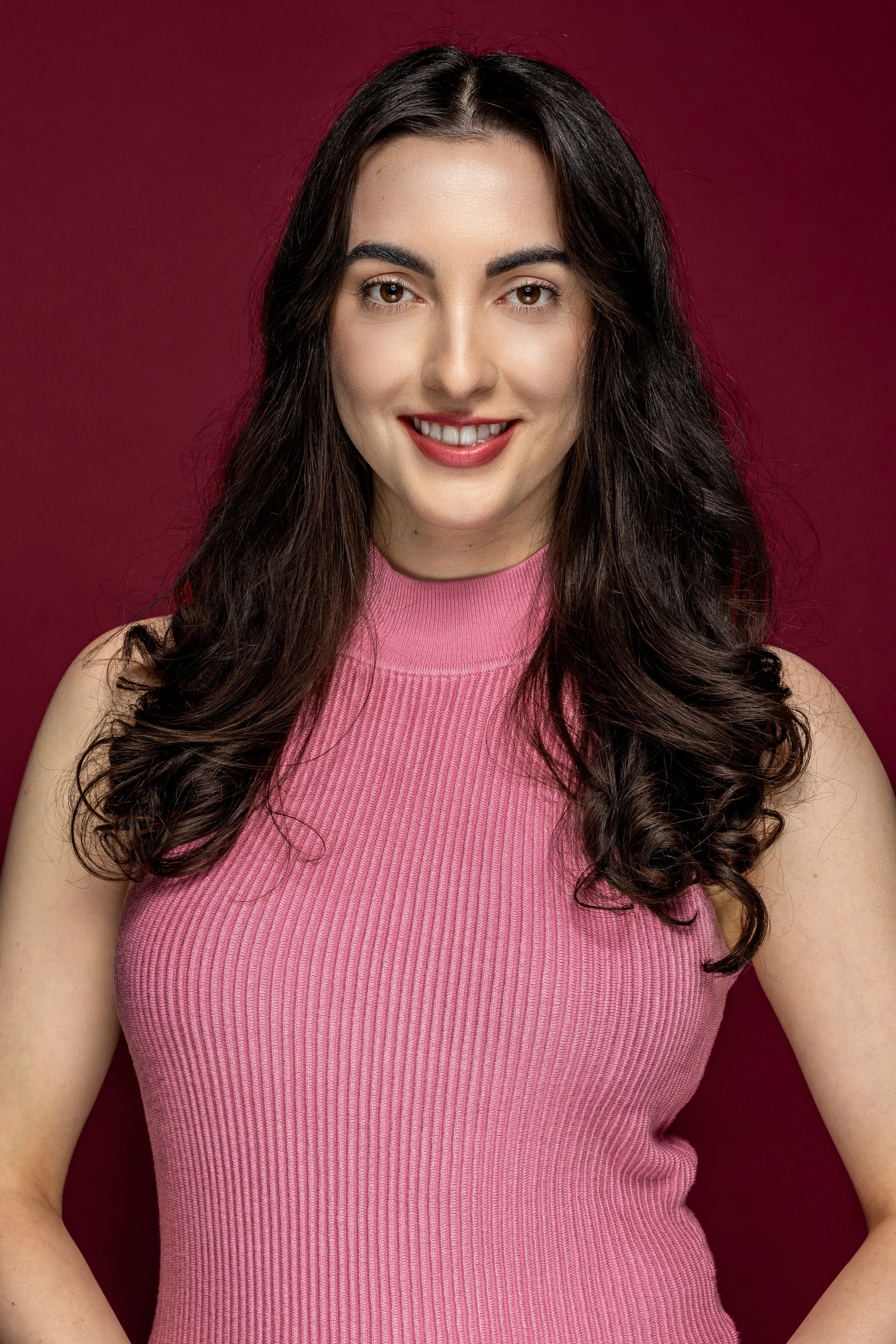 Portret van een jonge vrouw met lang, donkerbruin haar in een roze geribbelde top, lachend op een rode achtergrond.
