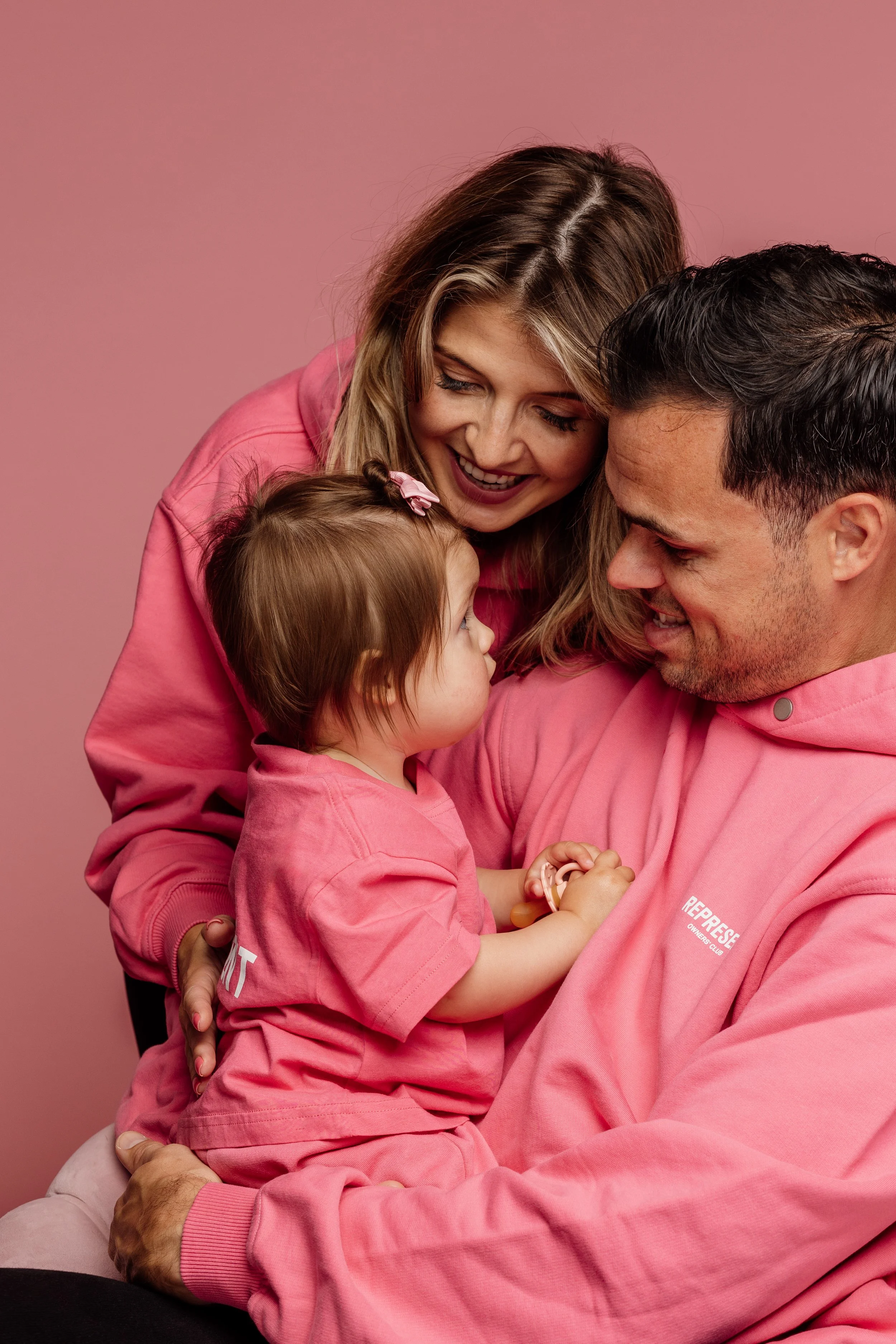Gezin bestaande uit een man, een vrouw en een klein meisje met dezelfde roze outfits, knuffelen en kijken naar elkaar tegen een roze achtergrond.