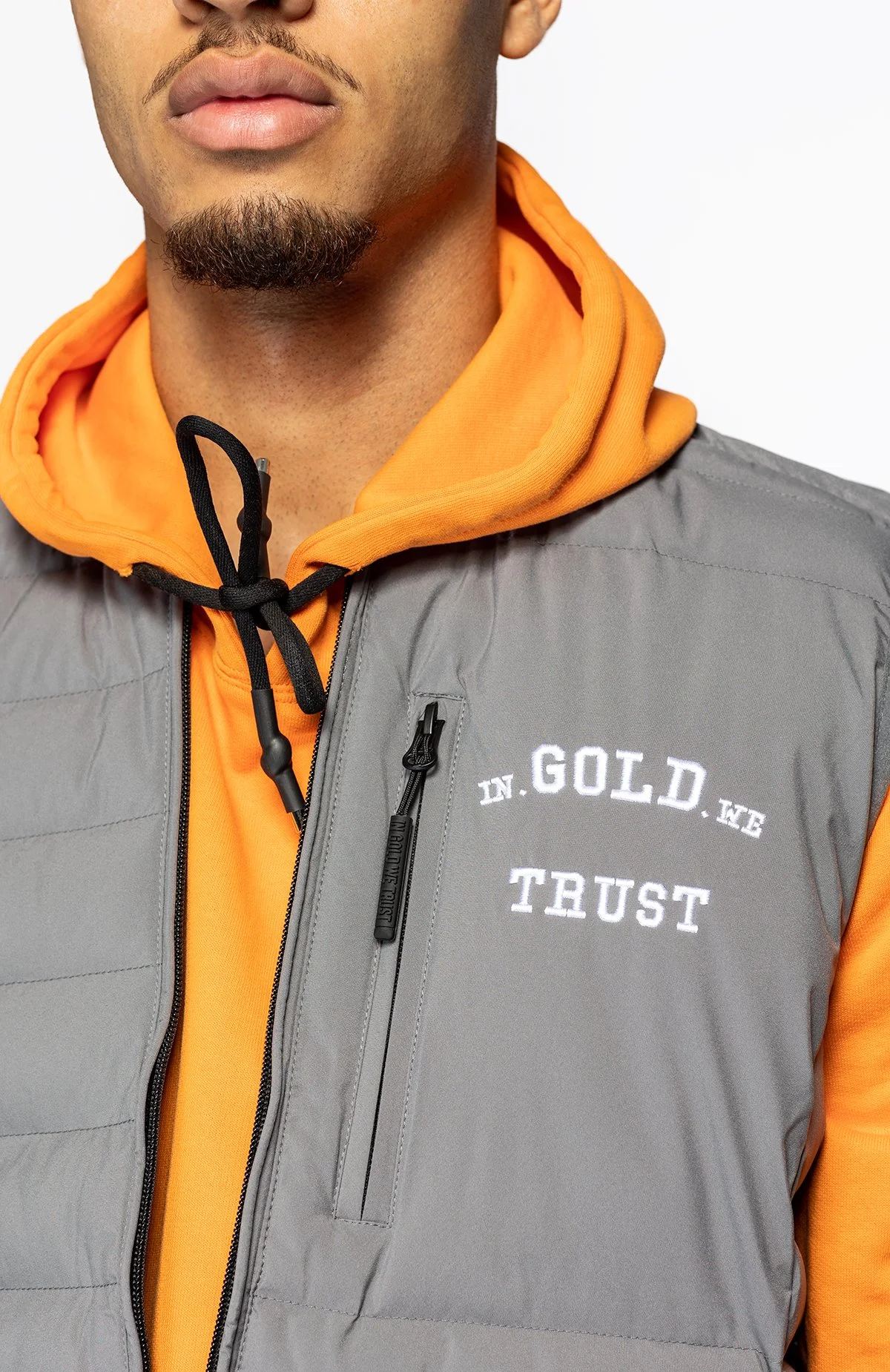 Close-up van een man die een geel hoodie en een grijze jas draagt met witte tekst 'IN GOLD WE TRUST' op de jas.