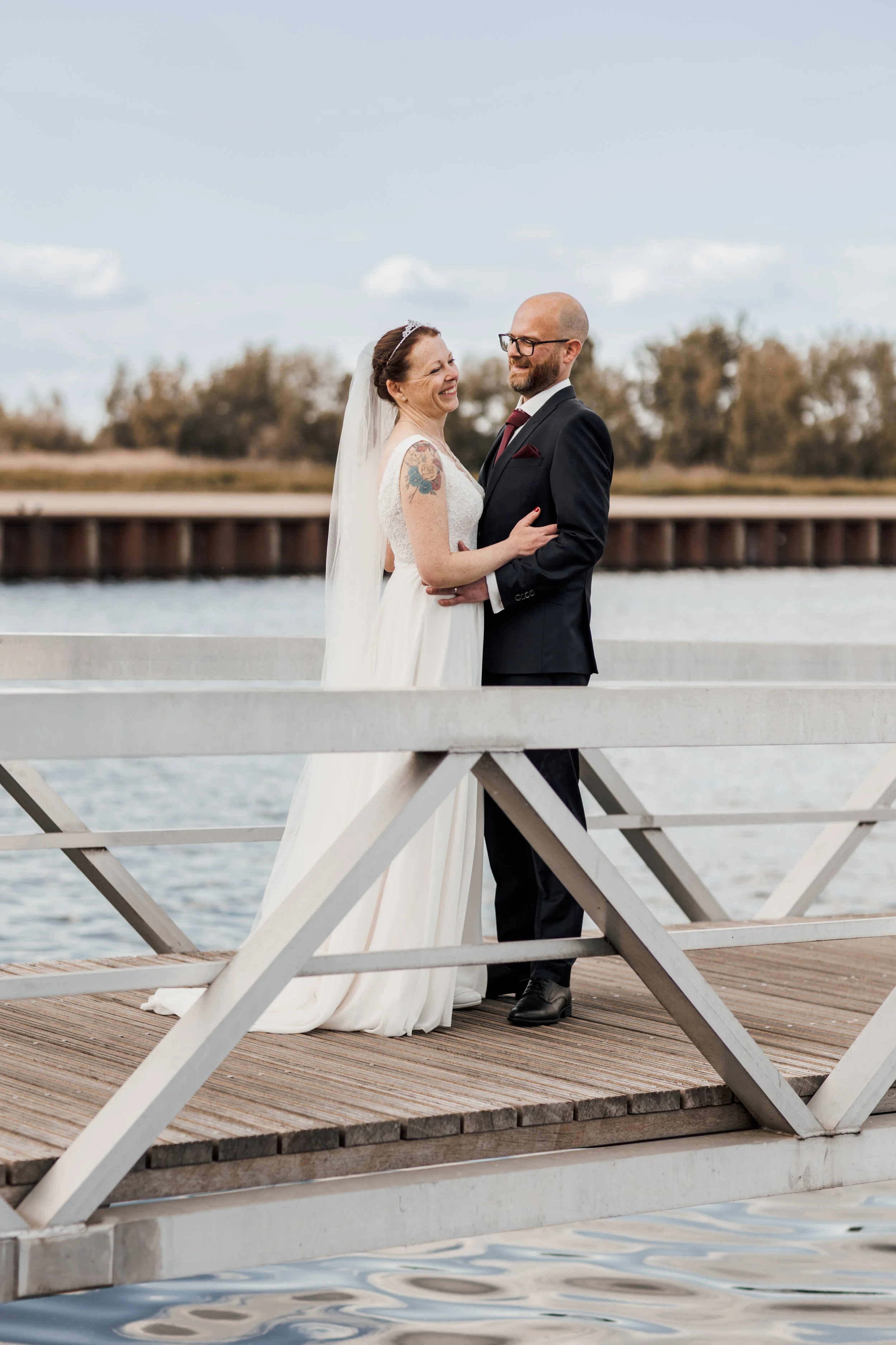 Een getrouwd paar op een houten pier bij het water, lachend en in elkaars armen, tijdens hun bruiloft.