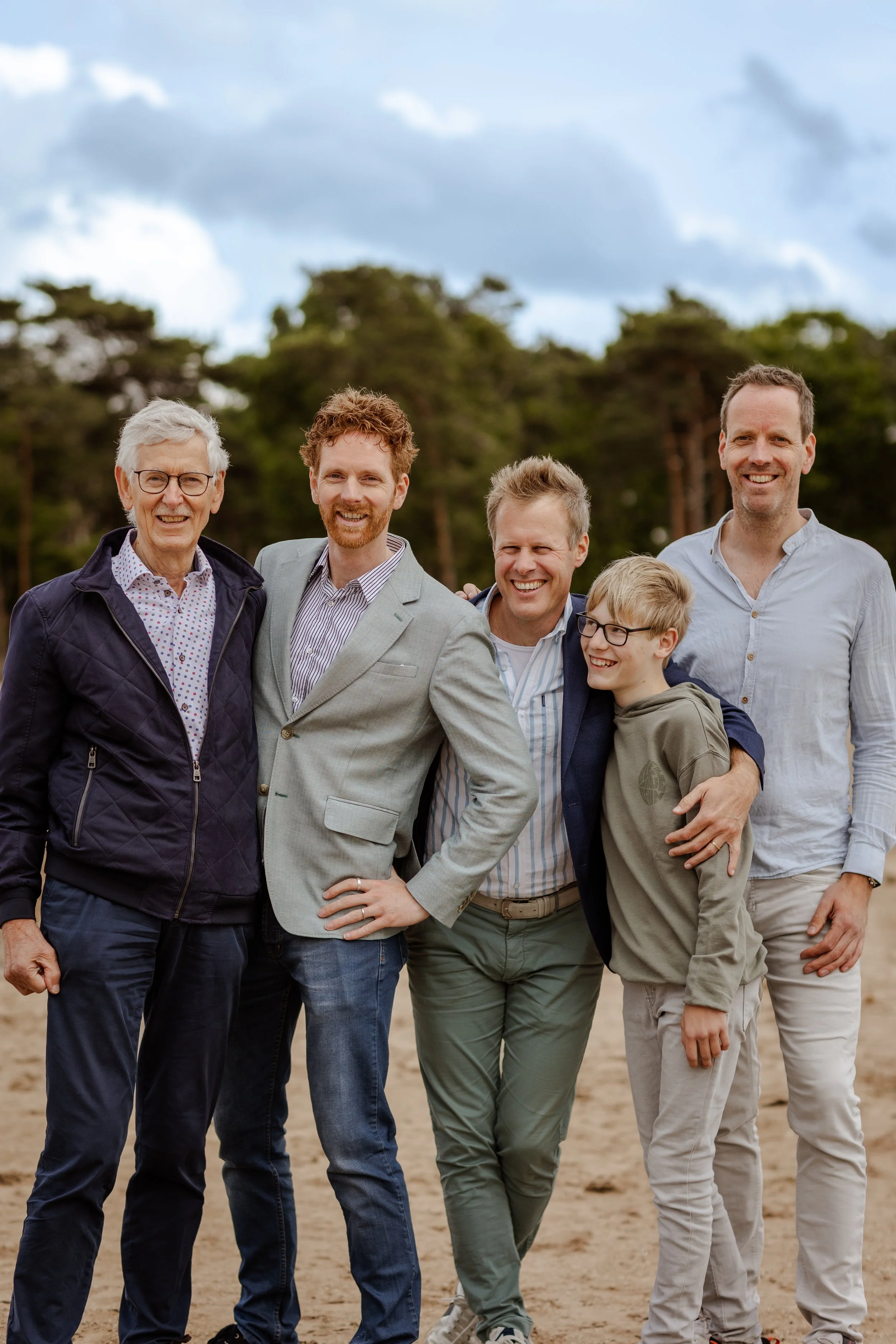 Vijf mensen staan samen op het strand, lachend en in een groep, met bomen en een bewolkte hemel op de achtergrond.