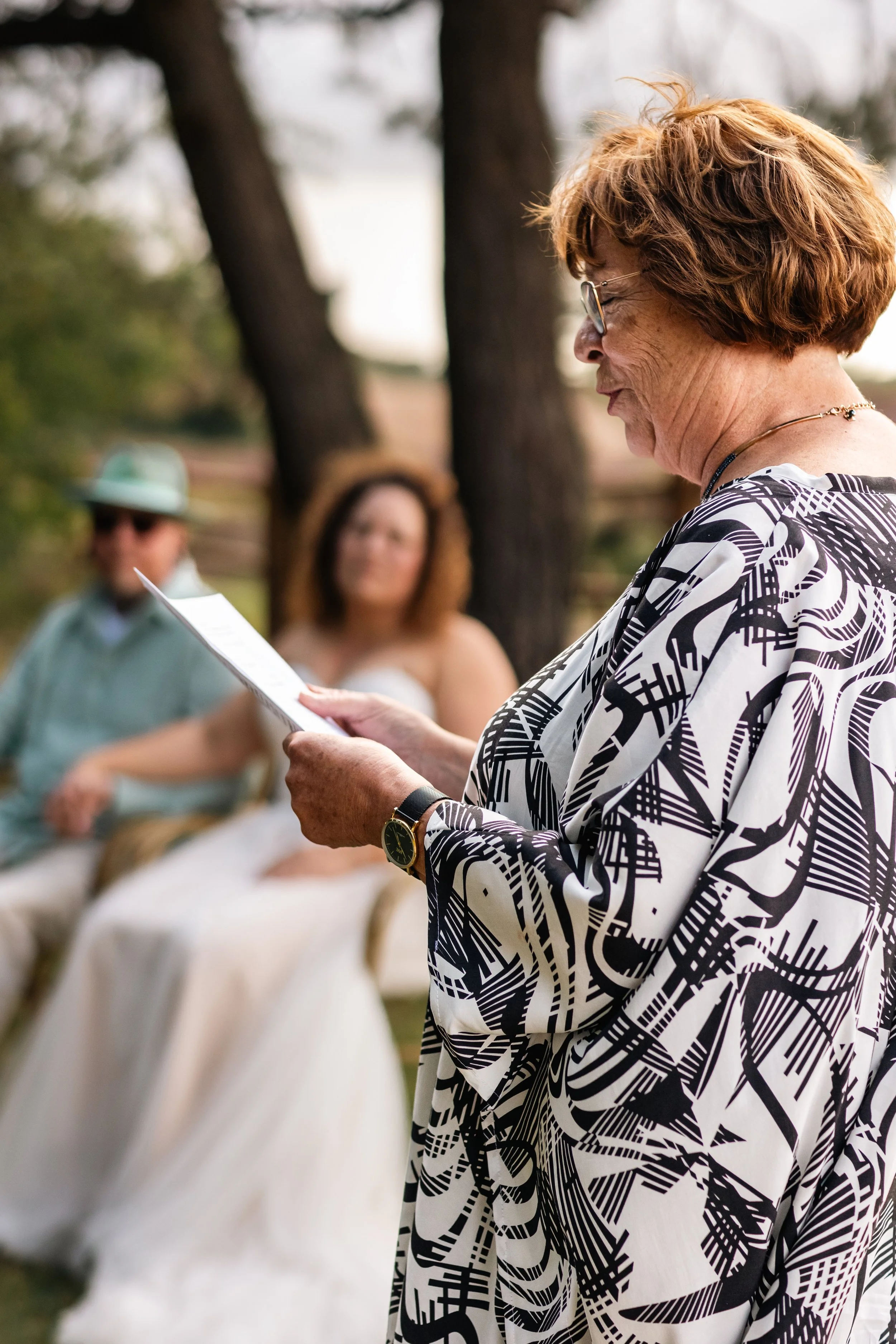 Een oudere vrouw leest een speech voor tijdens een buitenbruiloft, terwijl twee mensen op de achtergrond luisteren.