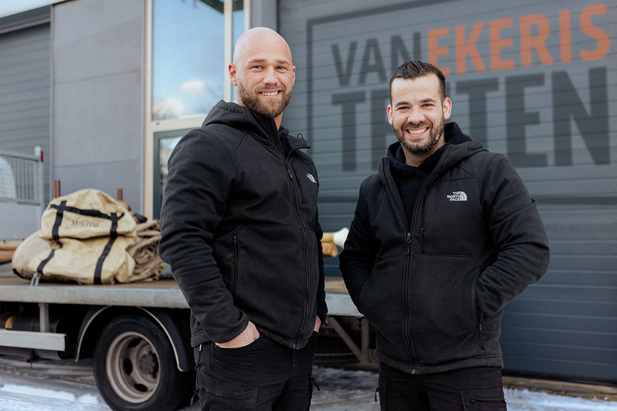 Twee mannen in zwarte jassen staan buiten voor een winkel. Ze glimlachen en poseer voor de camera.