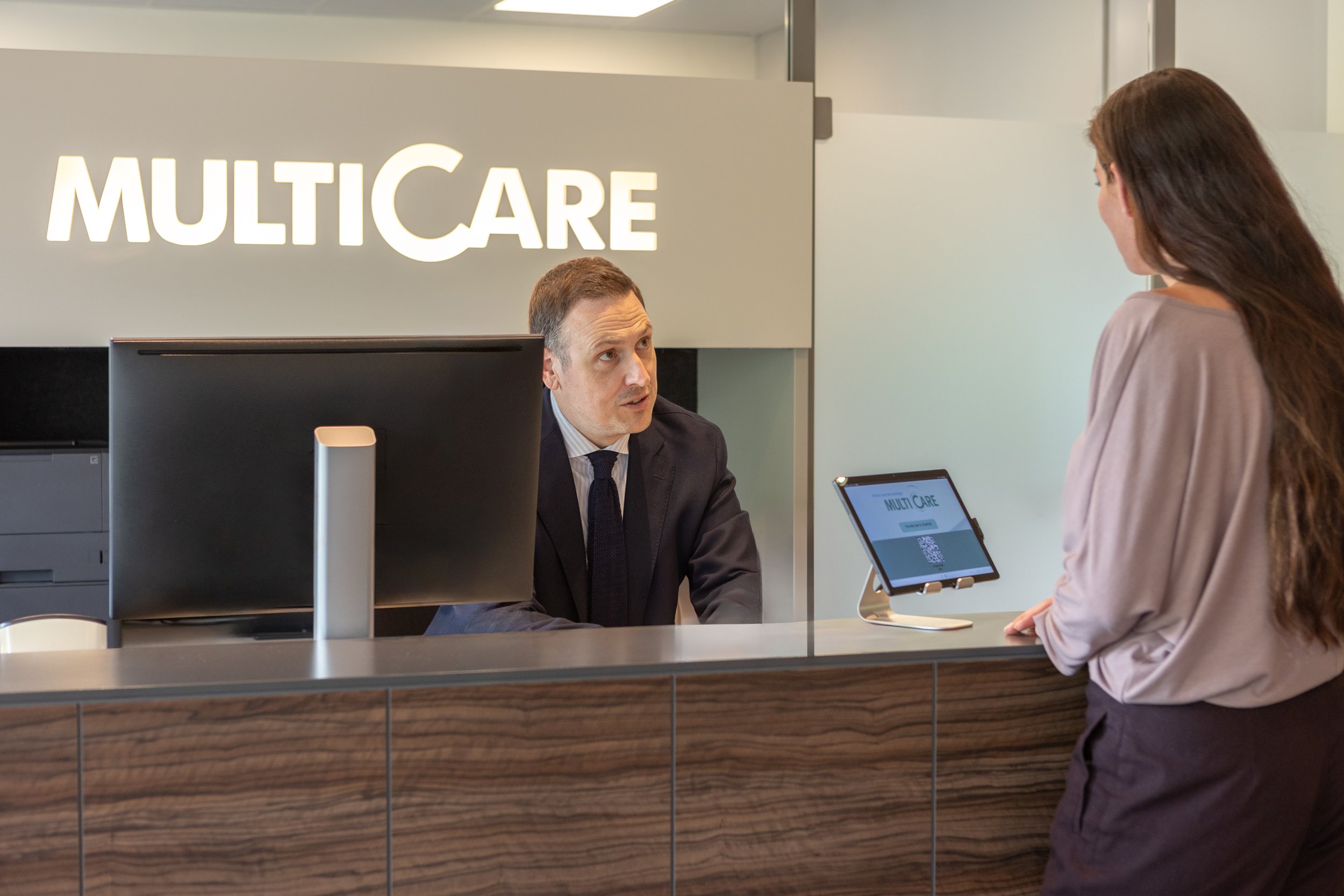 Man achter balie spreekt met vrouw in kantoor- of hotelomgeving met 'MULTICARE' sign.