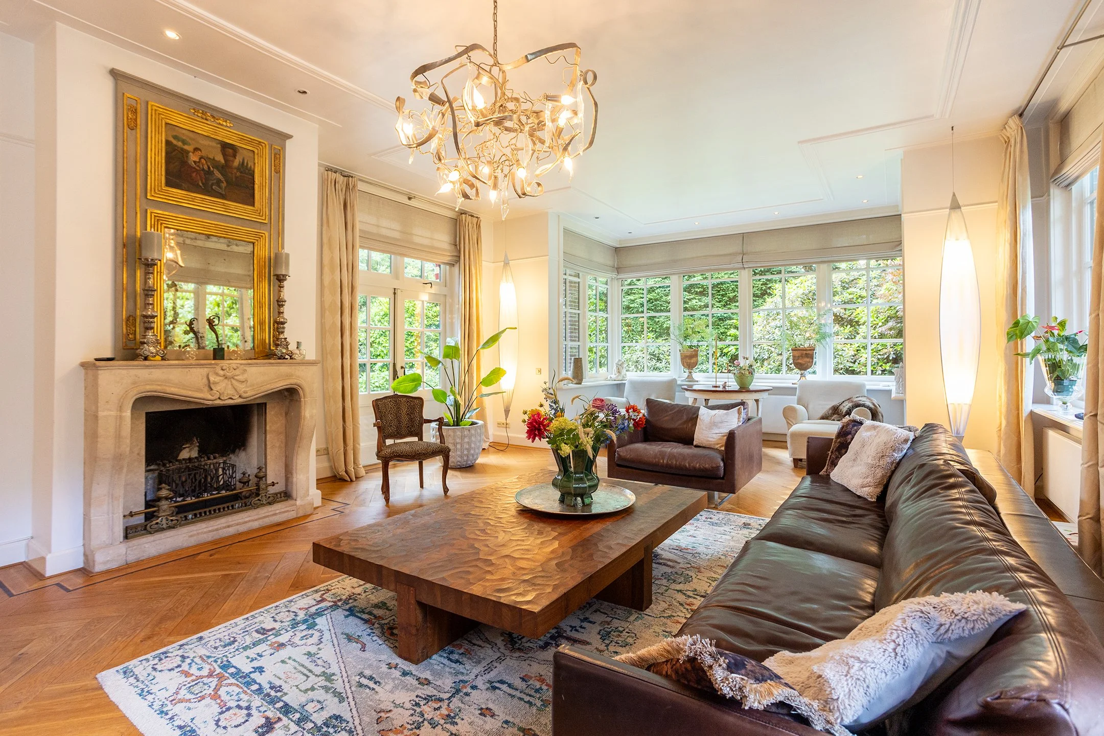 Een gezellige woonkamer met een open haard, grote ramen met uitzicht op groen, een chique chandelier, zachte meubels en decoraties en veel natuurlijk licht.