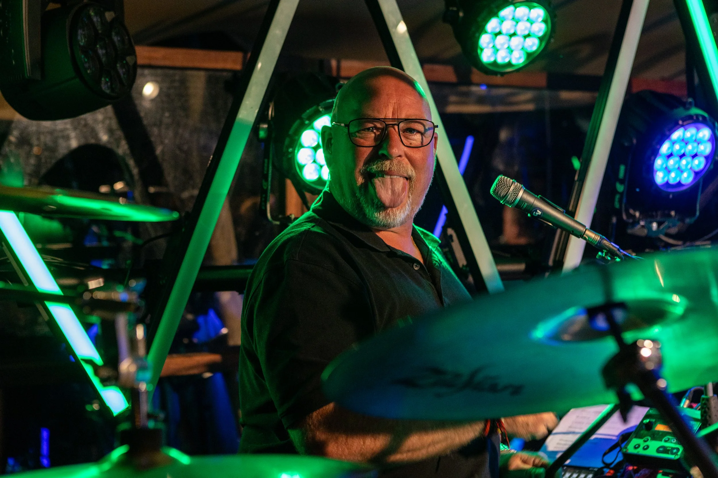 Man met een grijze baard, bril, lachend en met tongue out, speelt op drums onder groene en blauwe lichtstralen.