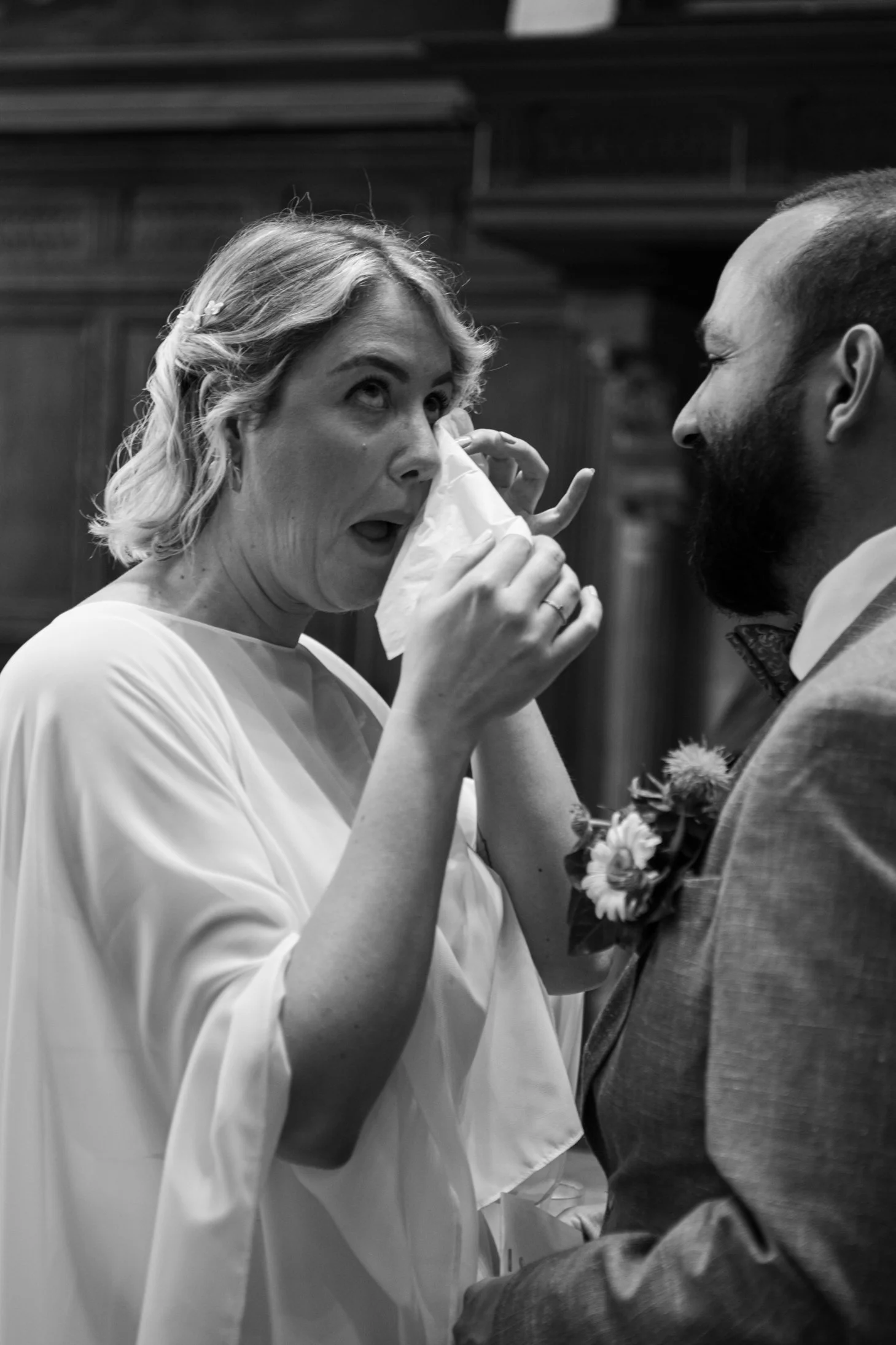 Een vrouw die een tissue gebruikt terwijl ze emotioneel is, en een man die naar haar kijkt, beiden in een trouwkleding, waarschijnlijk tijdens een bruiloft in een kerk.