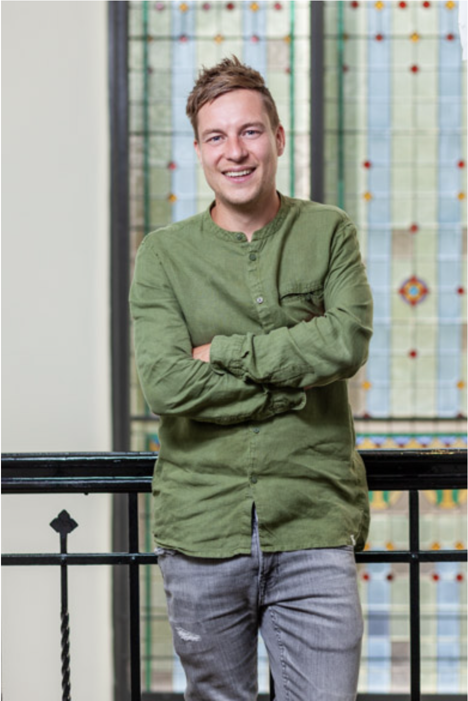 Lachende jonge man met groene blouse en grijze jeans, staand bij een zwart metalen railing met kleurrijk glas-in-lood raam op de achtergrond.