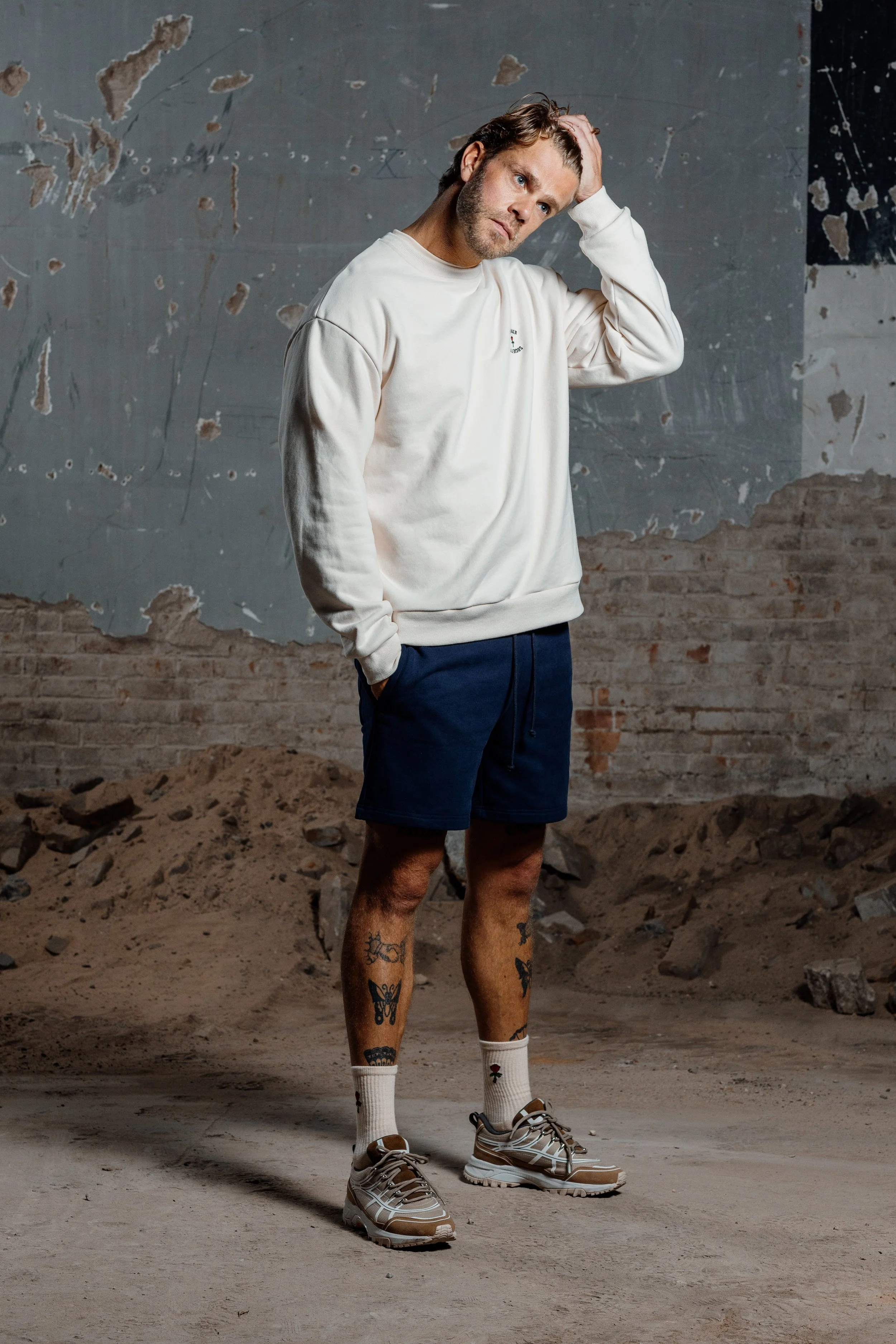 Een man in een witte sweatshirt, blauwe shorts, witte sokken, en sneakers, staat in een ruïne-achtige omgeving met een kapotte muur en stoffige vloer.