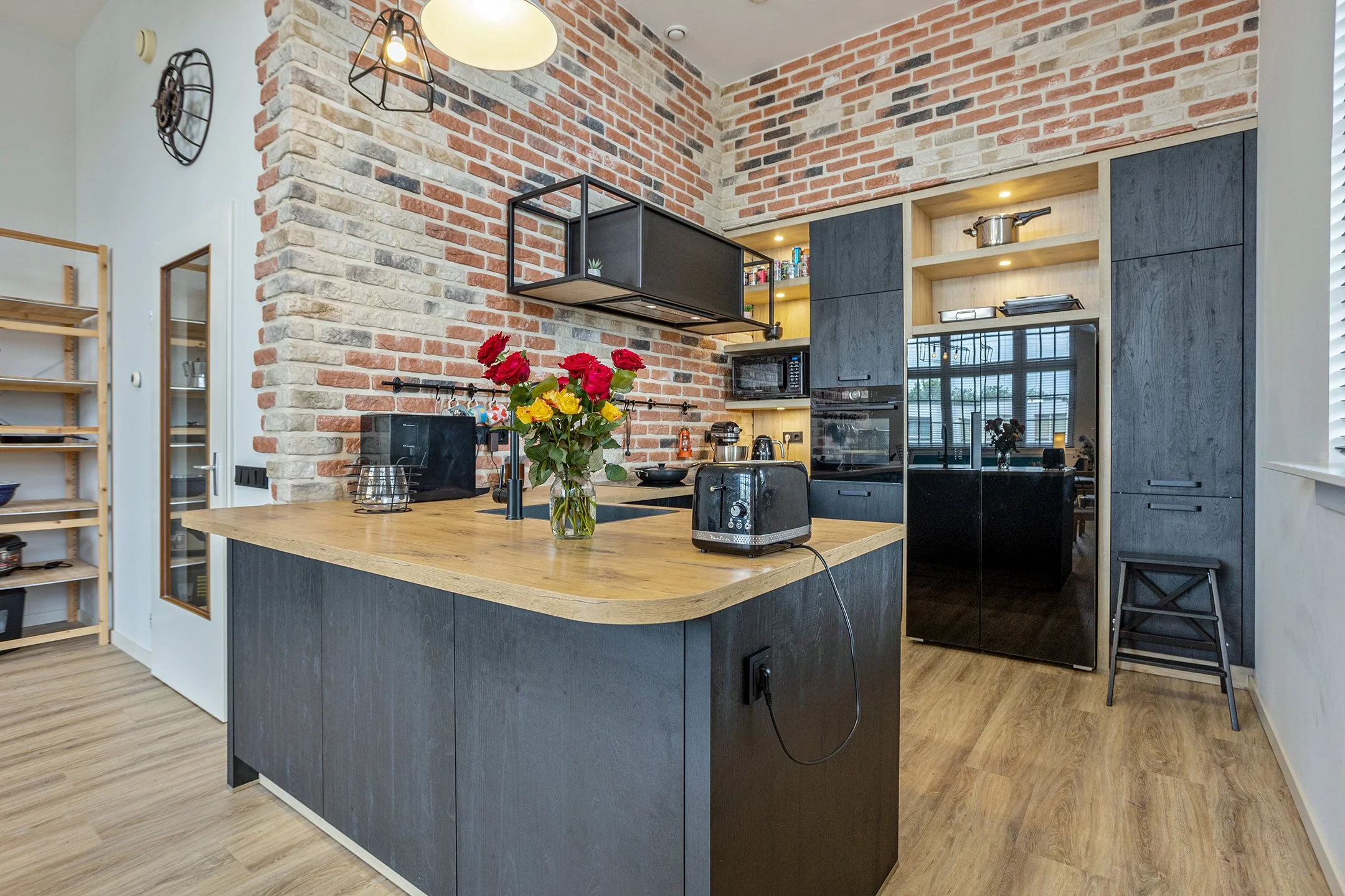 Moderne keuken met een houten eiland en zwart kastwerk, bakstenen muur, bloemen in een vaas, magnetron, broodrooster, en oven