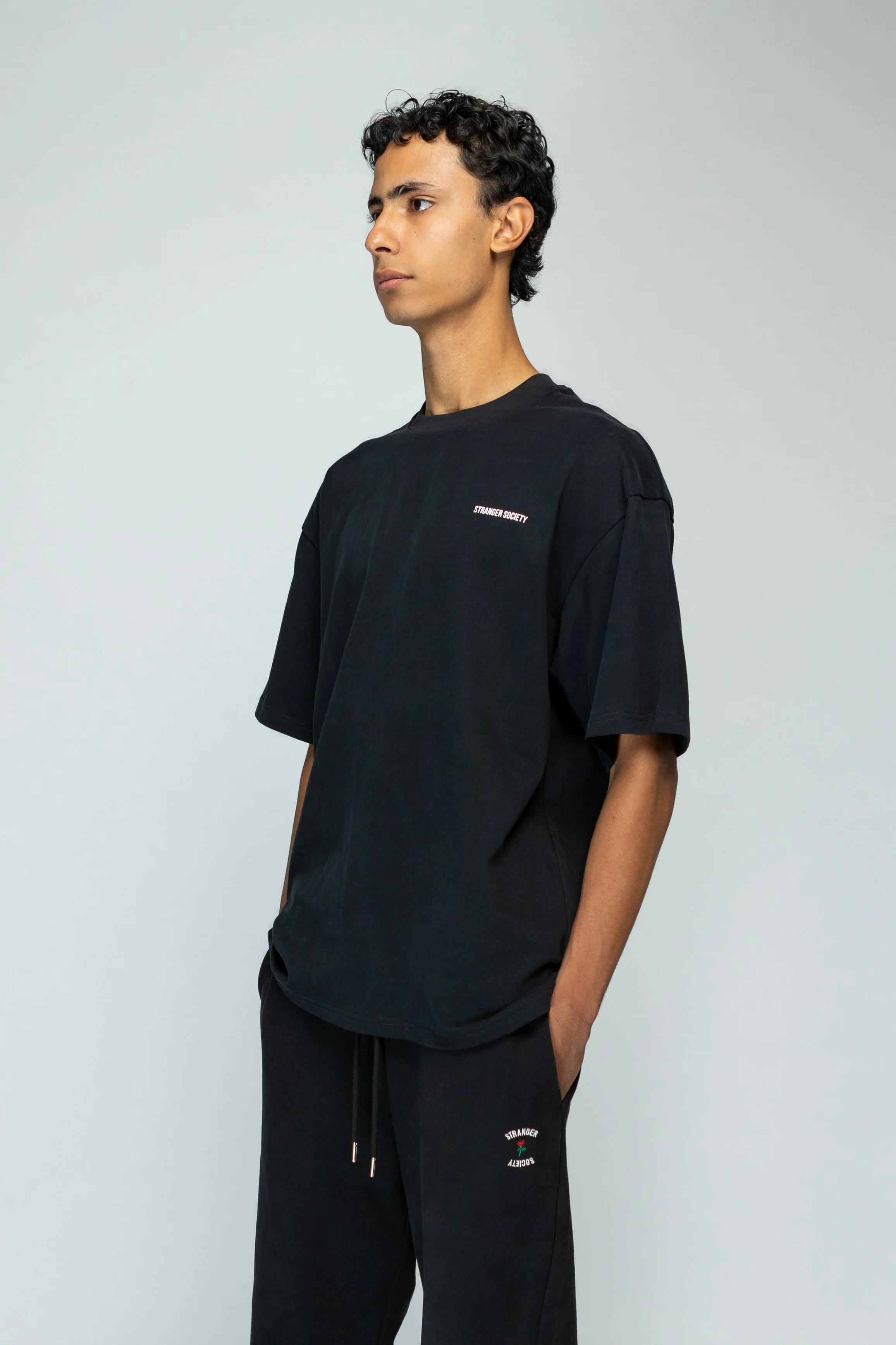 Jonge man met kort, krullend haar in een zwart T-shirt en joggingbroek met teksten en iconen, staand met handen in zakken, tegen een lichte effen achtergrond.