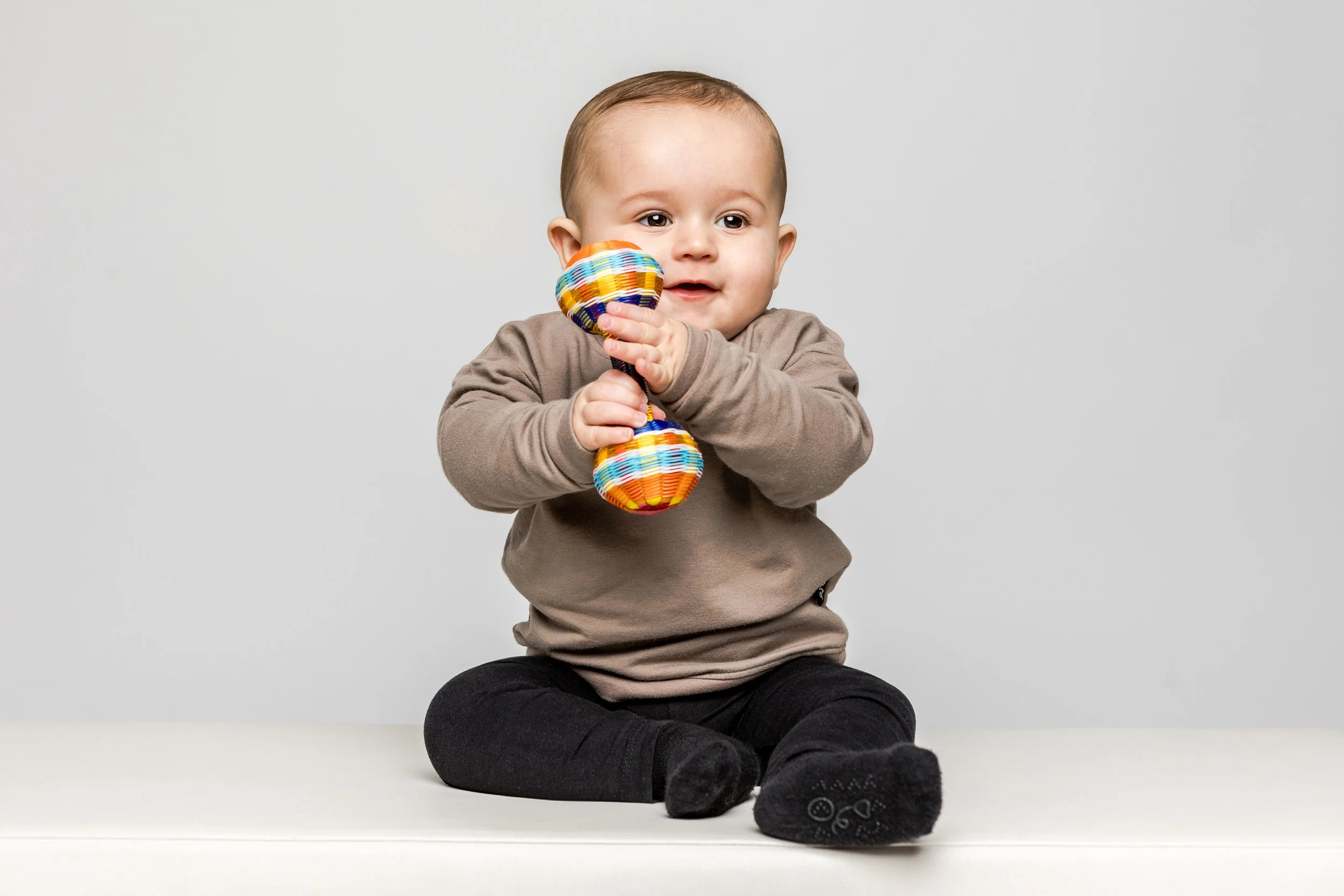 Een schattige baby die op een witte bank zit, een gekleurd rammelaar vasthoudt en geïnteresseerd kijkt.