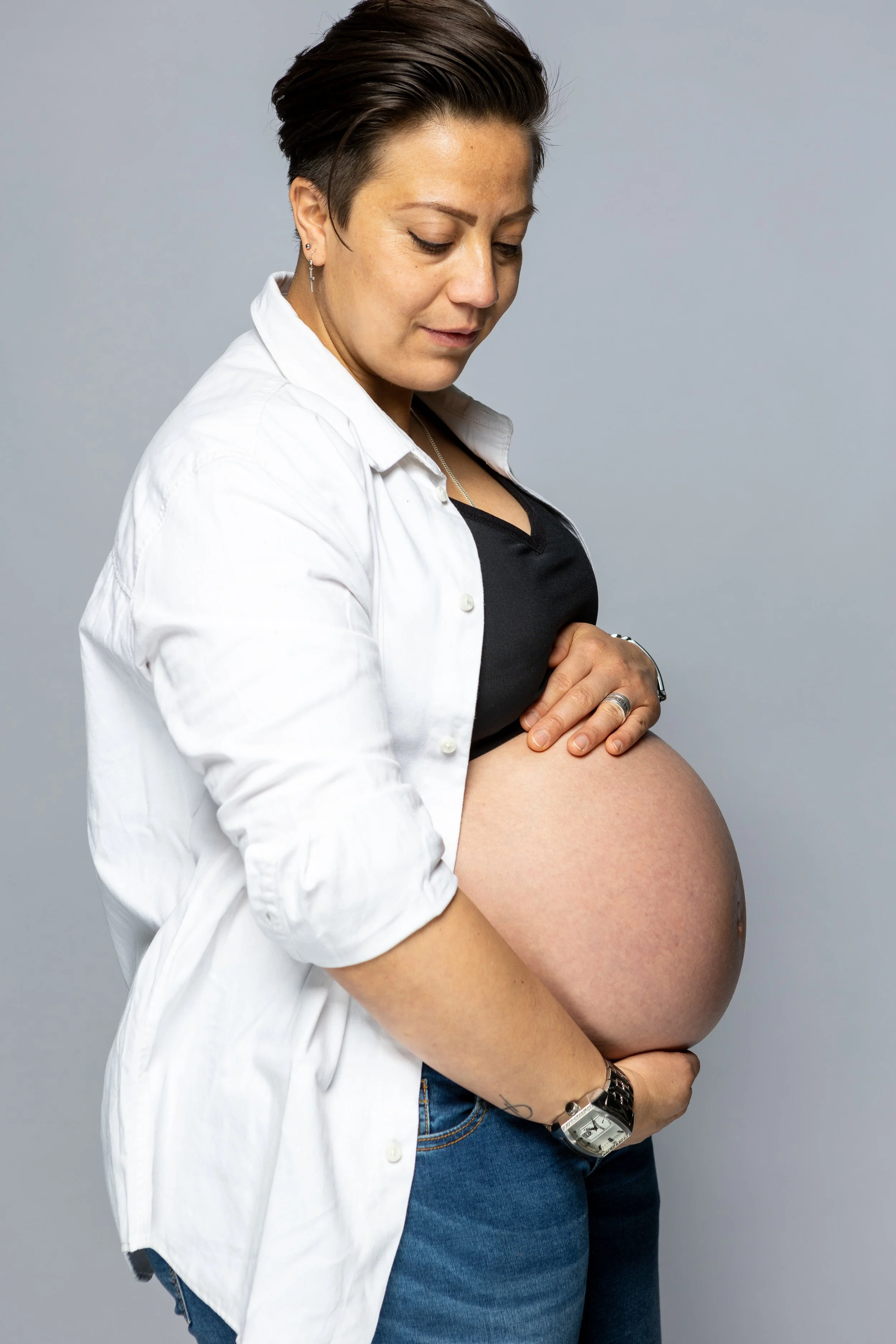 Een zwangere vrouw met korte, donkere haar draagt een zwarte top en een witte overhemd, terwijl ze haar buik vasthoudt en naar haar buik kijkt.