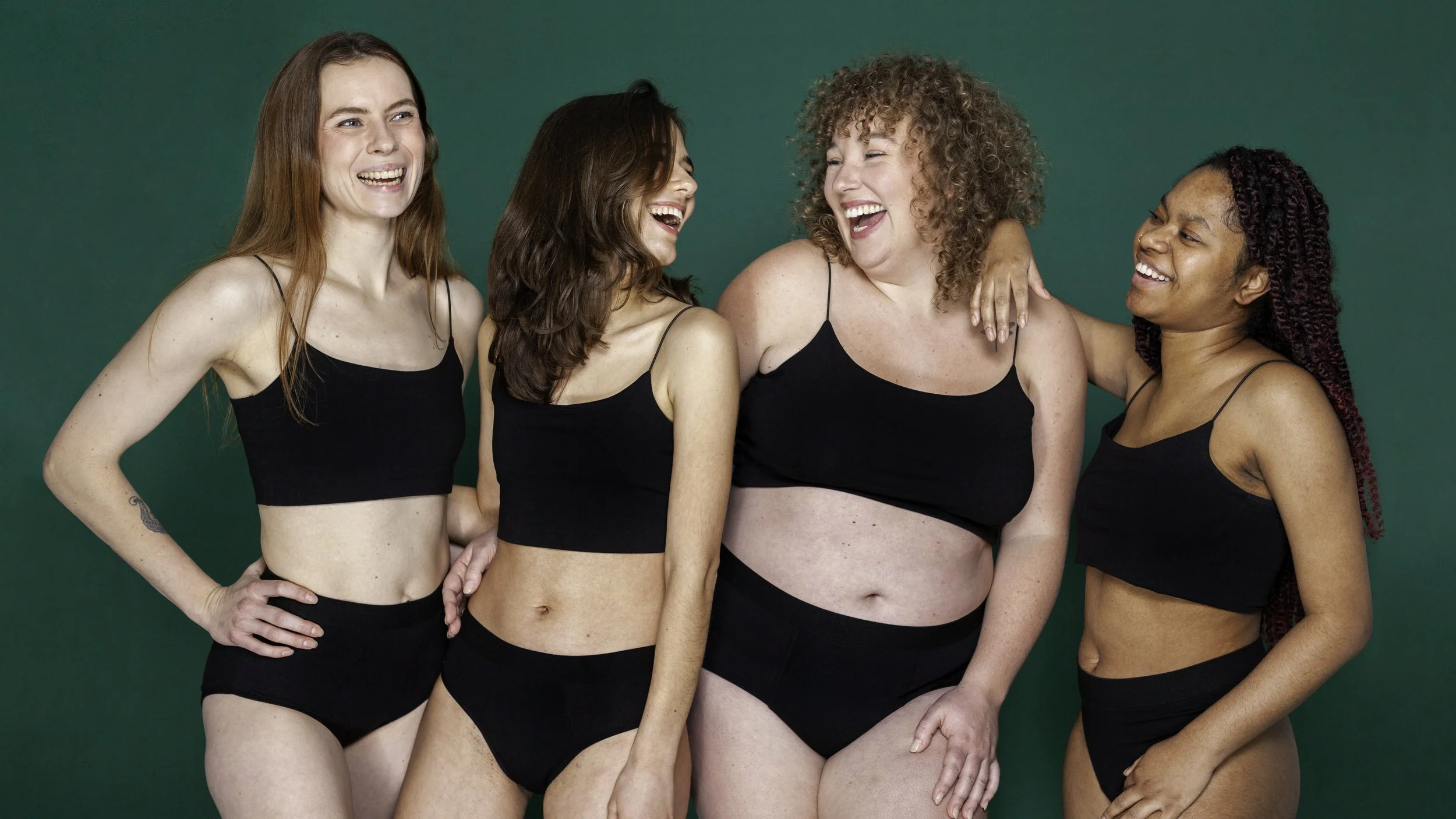 Vijf vrouwen lachen en hebben plezier samen, gekleed in zwarte onderkleding, tegen een groene achtergrond.