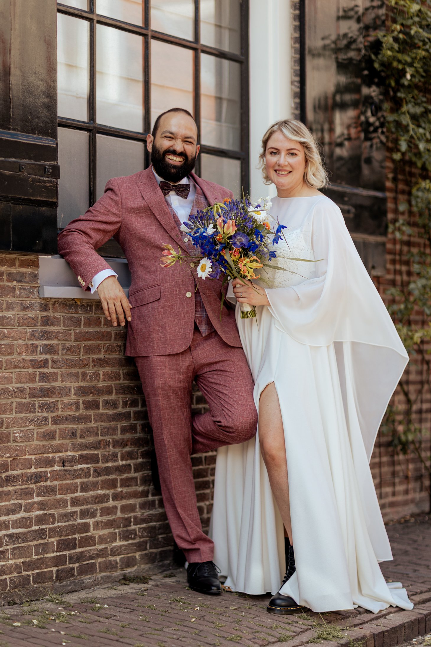 Een getrouwd stel poseert buiten, met de bruid die een boeket met blauwe, witte en roze bloemen vasthoudt en de bruidegom in een roestkleurig pak.