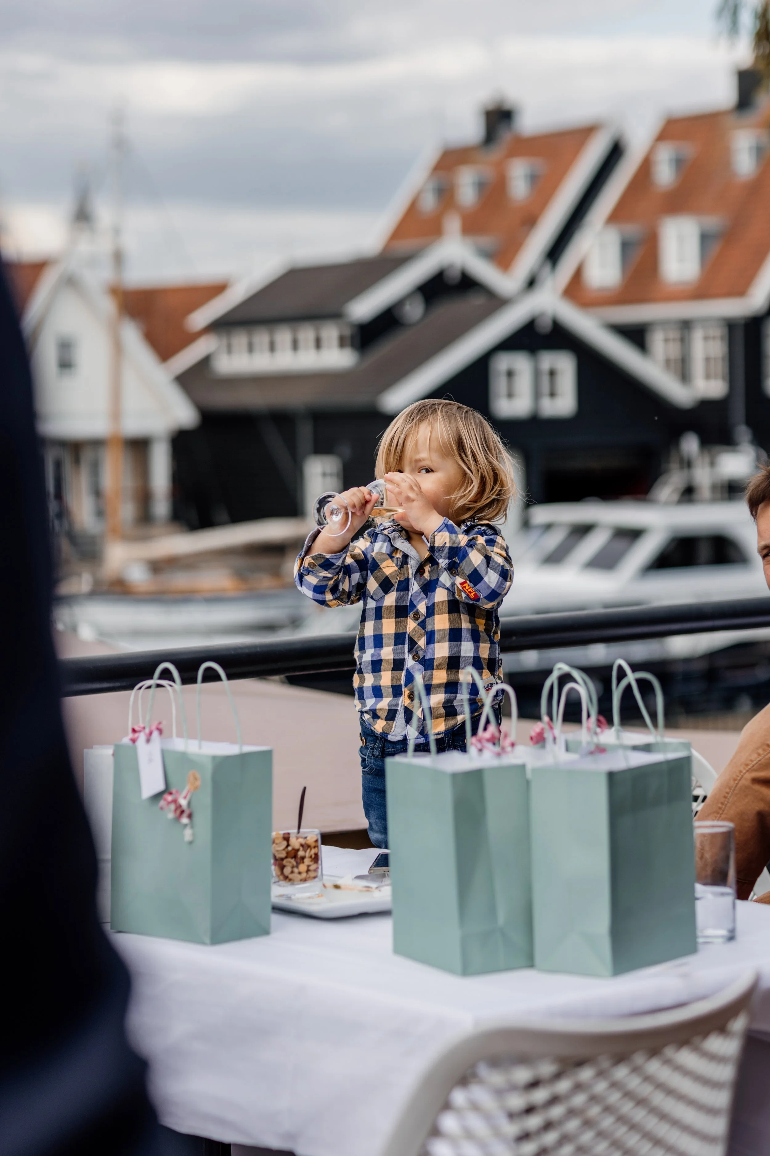 Een jonge jongen met gekleurde geruite blouse drinkt uit een wijnglas aan een tafel met cadeauzakken en snacks, buiten bij een haven.