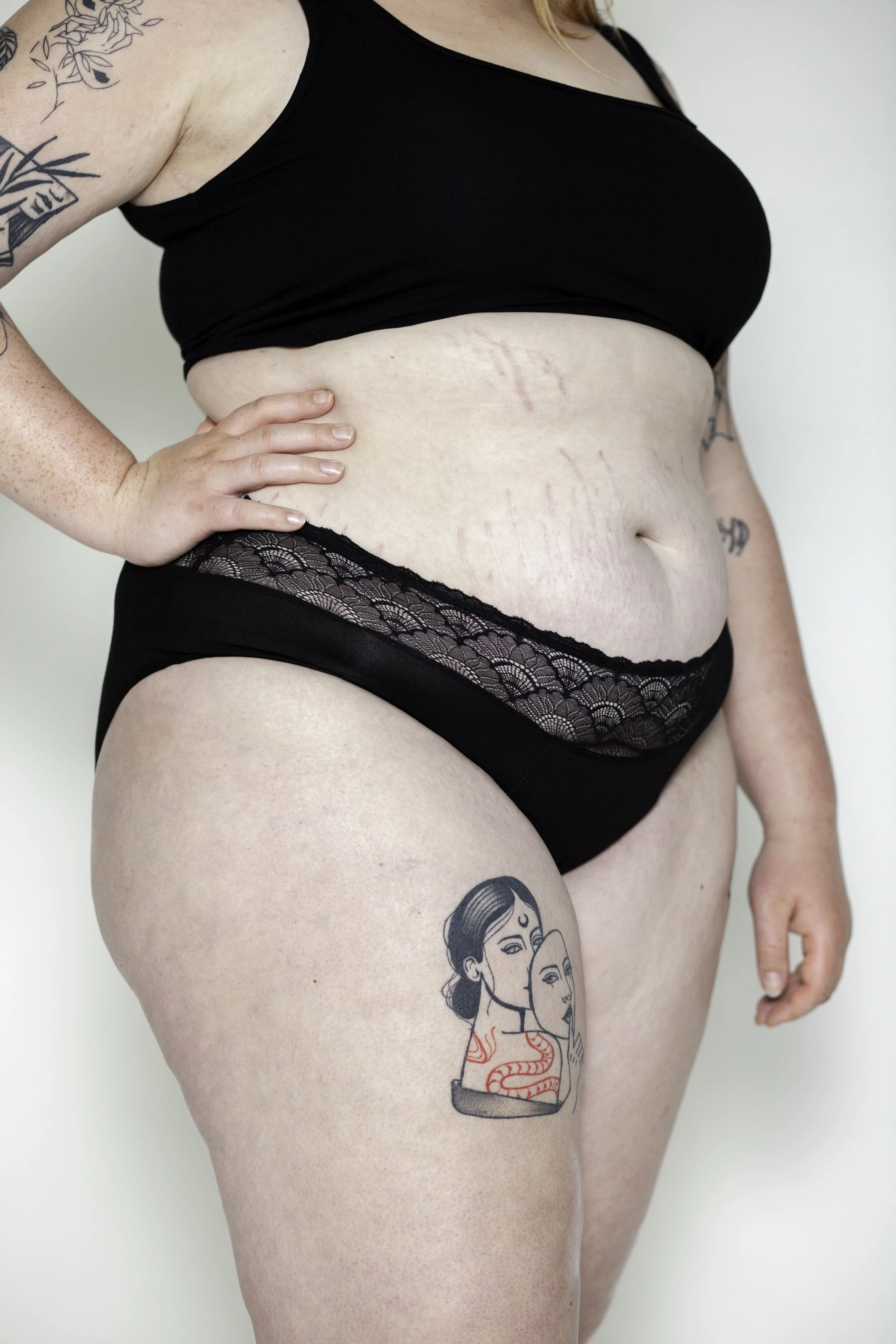 Draagt black ondergoed met lace detail, tatoeages op arm en dij; de tatoeage op de dij toont twee vrouwen met lange haar, een met een kroon op haar hoofd, en een slang om haar nek.