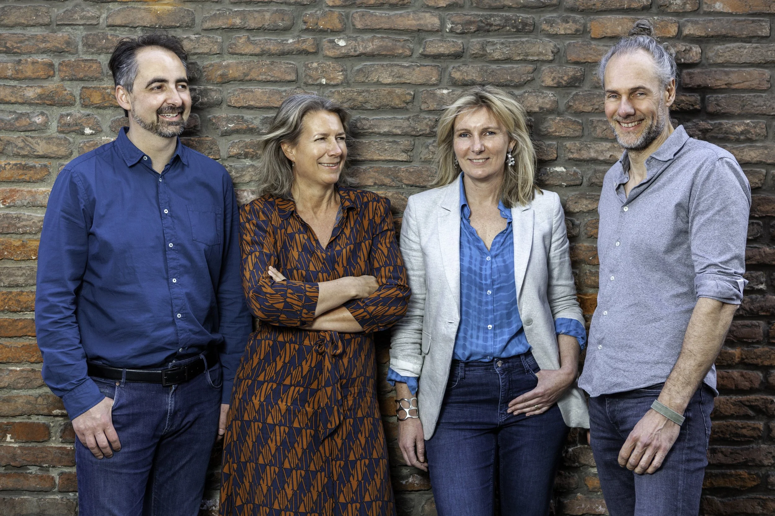 Vijf mensen staan samen voor een bakstenen muur, drie vrouwen en twee mannen, lachend en in geklede kleding.