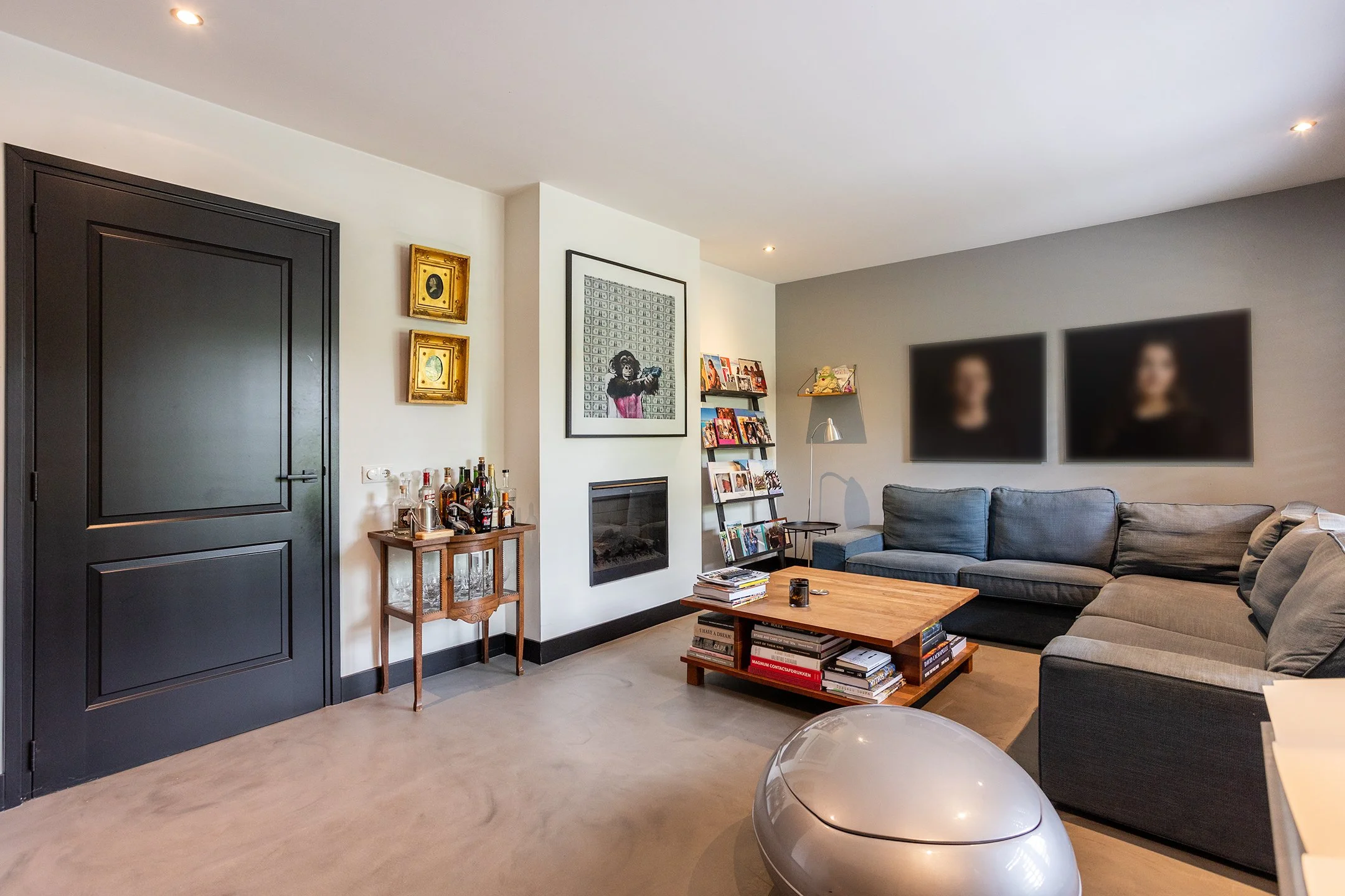 Moderne woonkamer met een grote L-vormige bank, een houten salontafel vol boeken, een tv met opgerolde blurrende beelden, een haard, een boekenrek, en een barwagen met drankflessen.