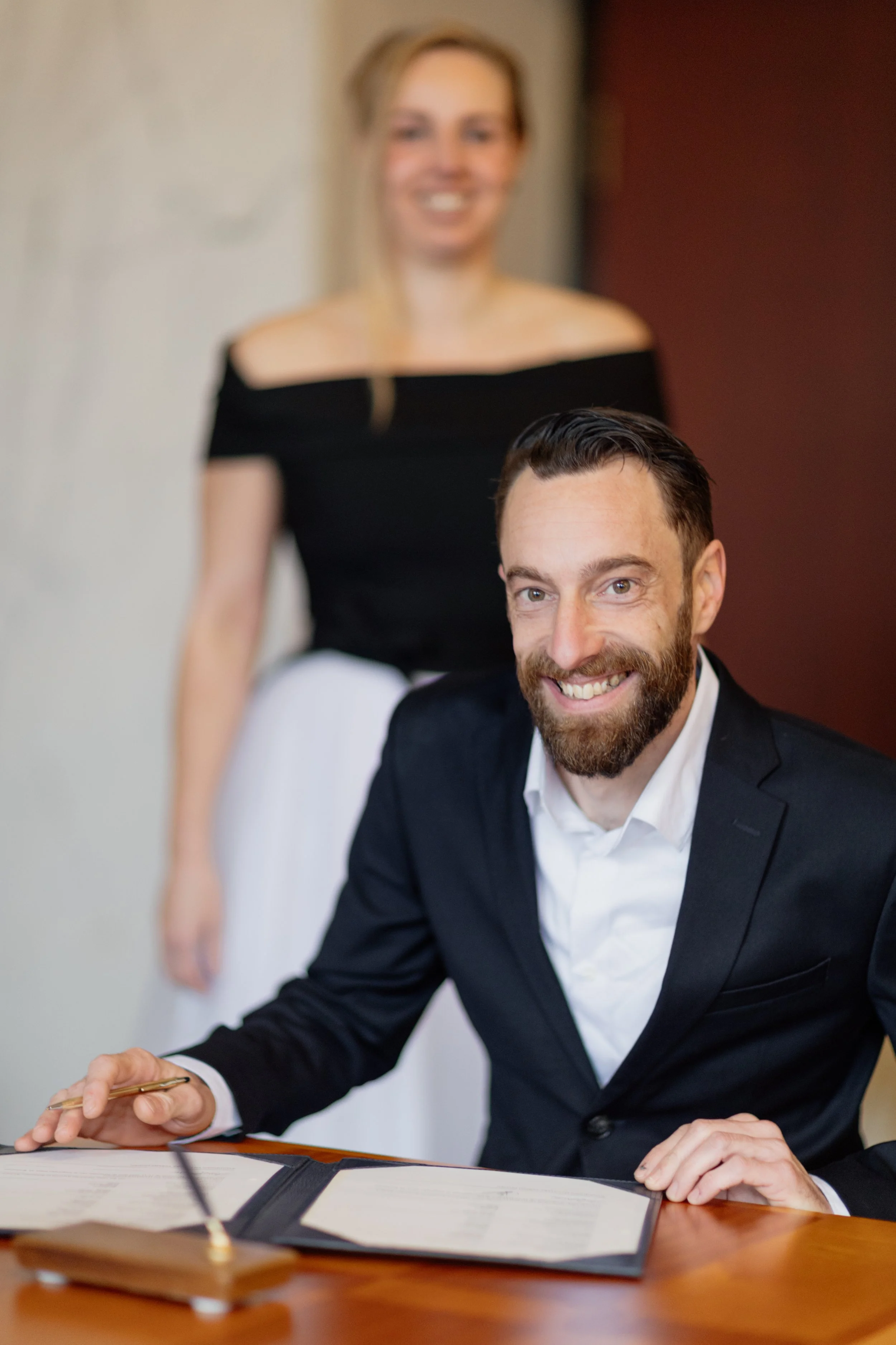 Man in een zwart pak die een getekend document ondertekent, achter hem vrouw in een zwarte top en witte rok, die glimlachend in de camera kijkt.