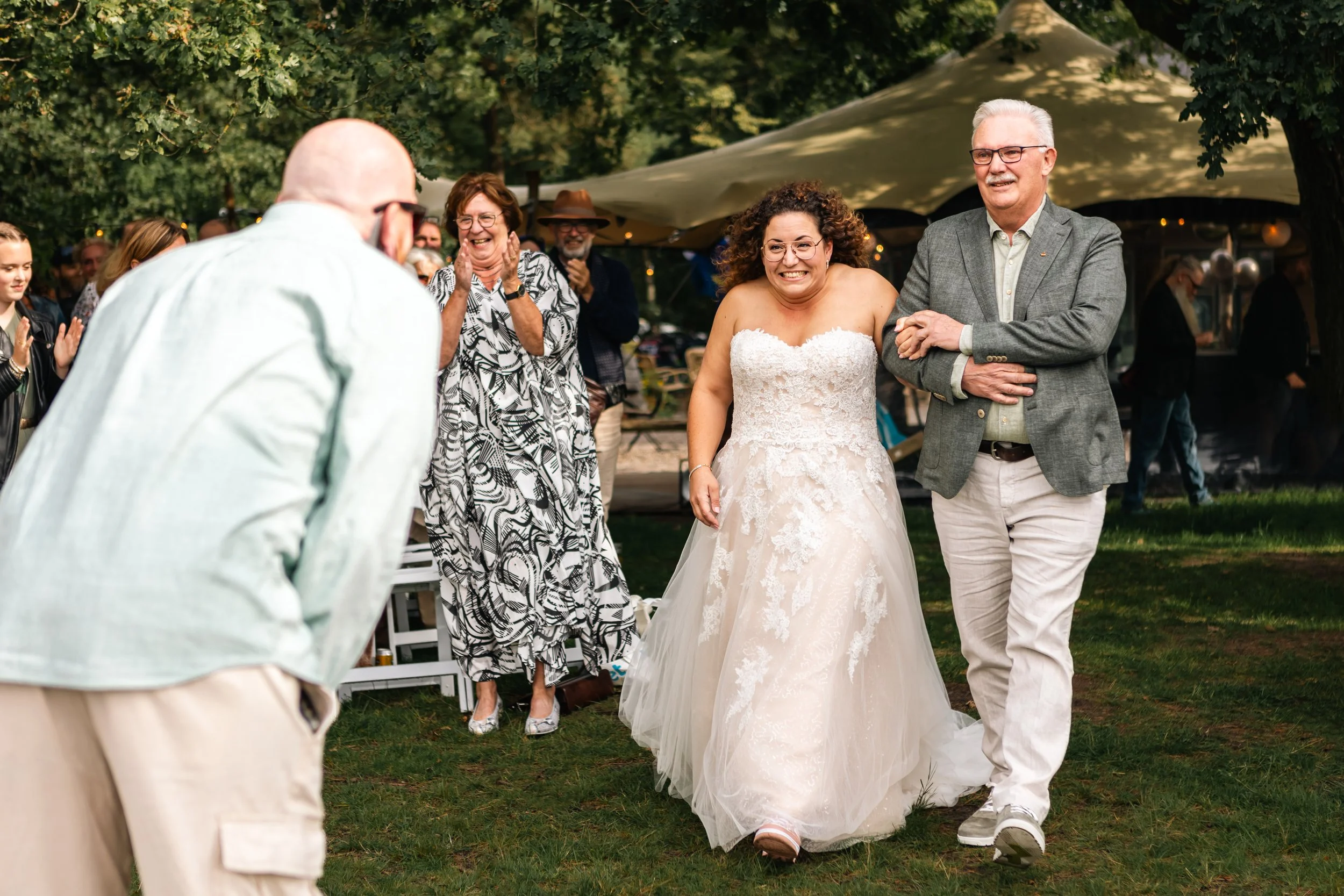 Bruidsmoeder en vader lopen samen lachend over gras tijdens een huwelijksfeest, omringd door feestgangers onder een boom en in een tent.