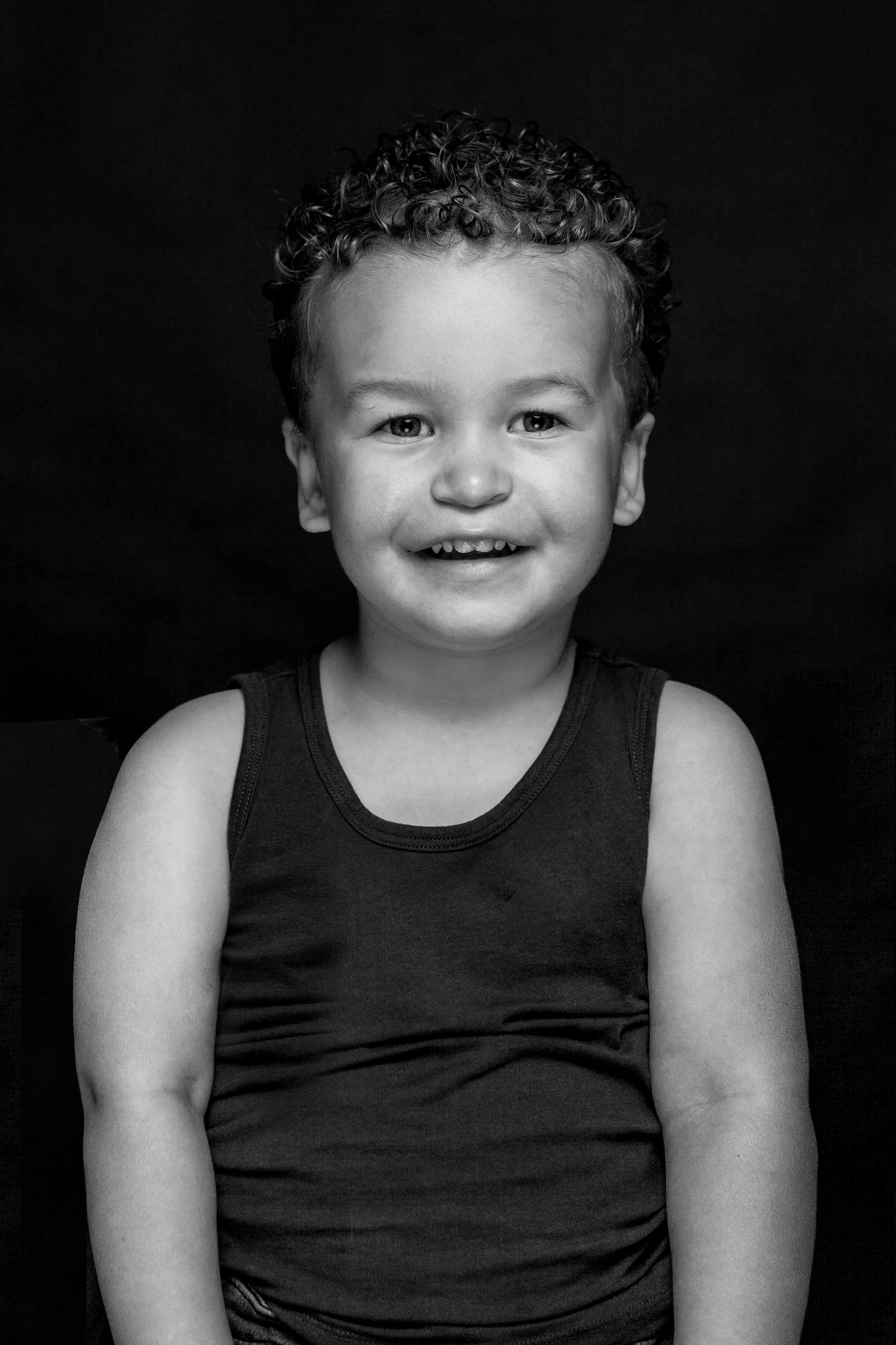 Zevenjarige jongen met donkere, krullende haar en een glimlach, zittend voor een zwarte achtergrond.