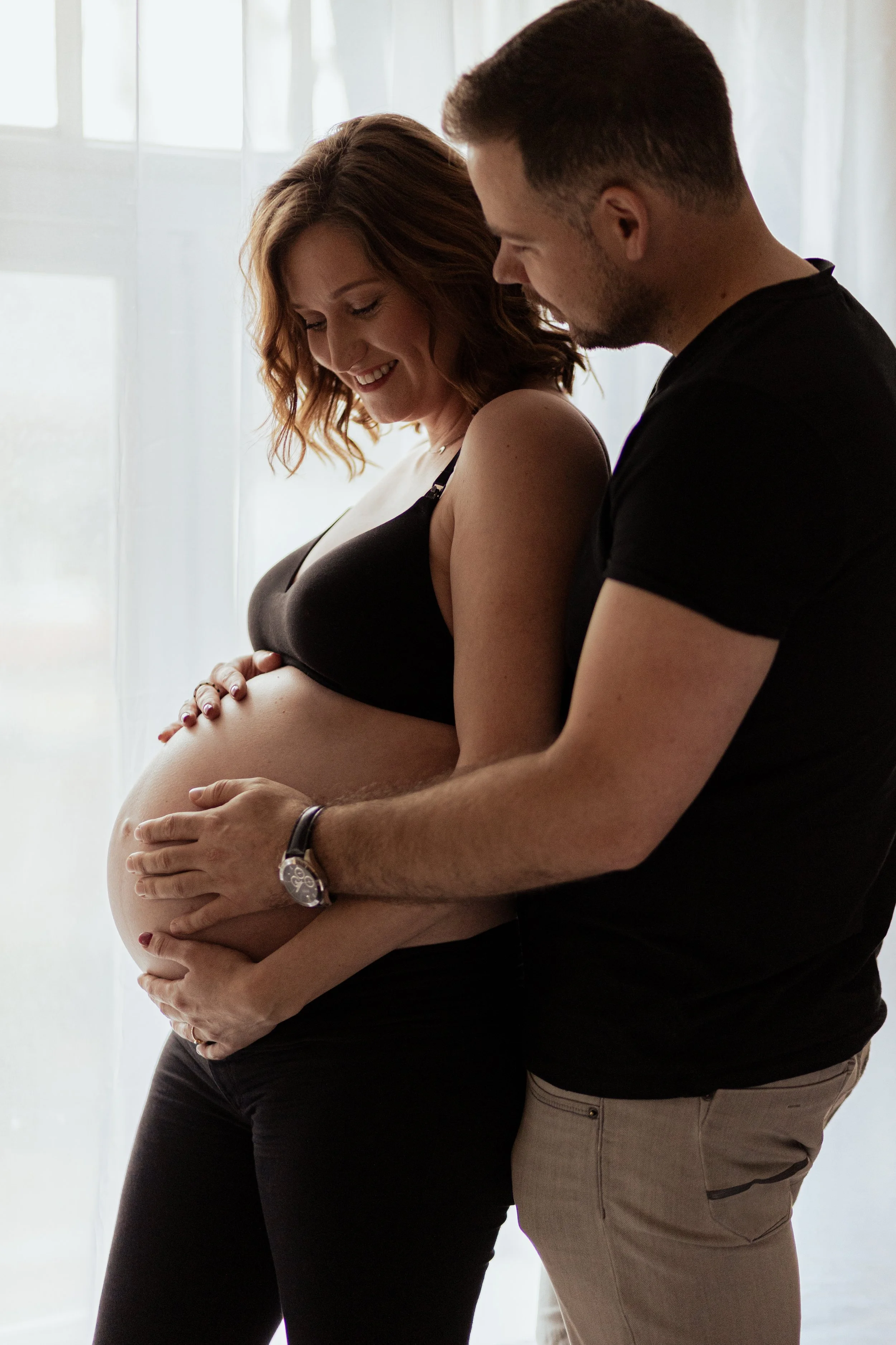 Een zwangere vrouw en haar partner die samen naar haar buik kijken. Ze staan voor een raam met witte gordijnen, de vrouw lacht en de man houdt haar buik vast.