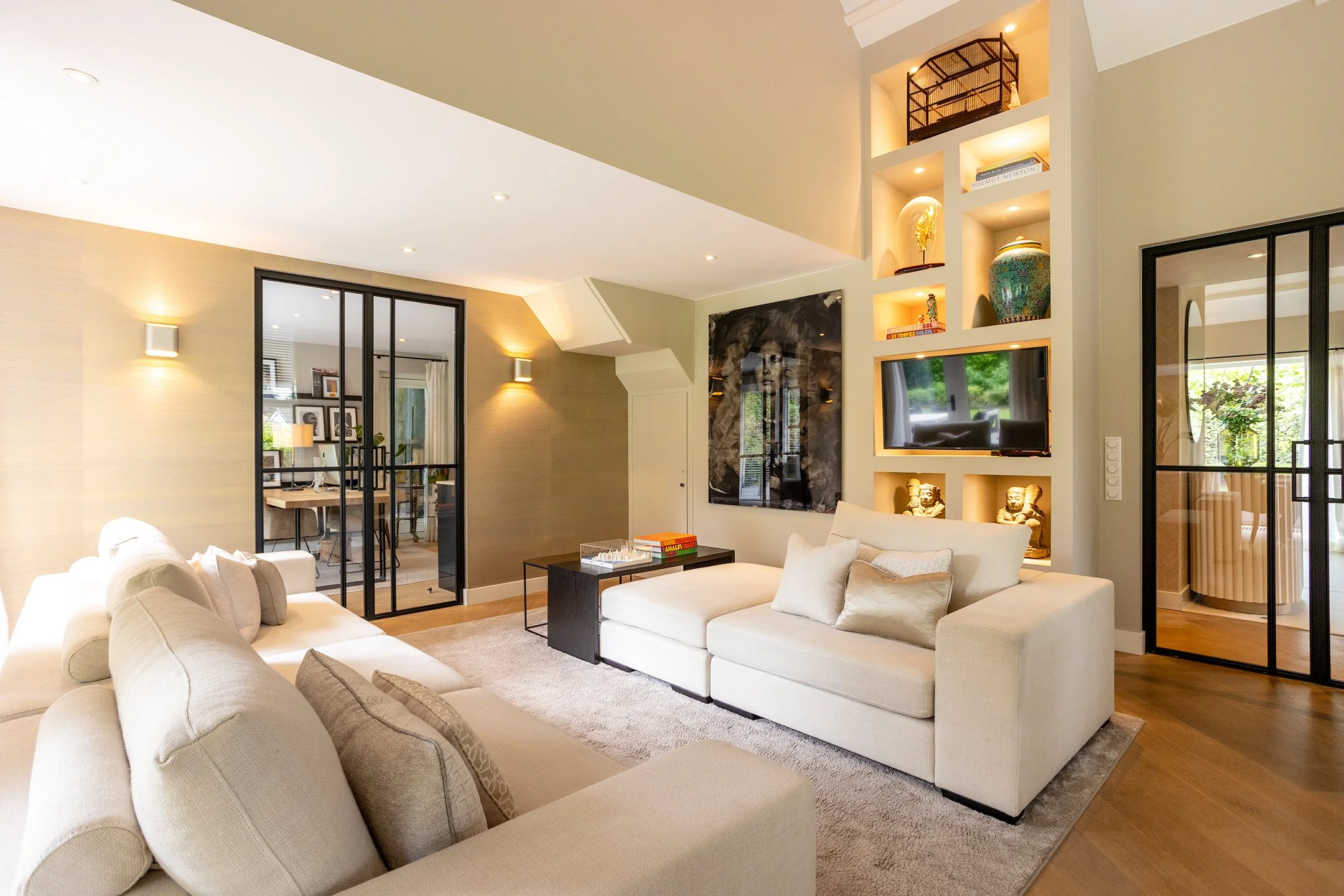 Modern woonkamer met beige banken, wanddecoratie, ingebouwde verlichting en glazen schuifdeuren.