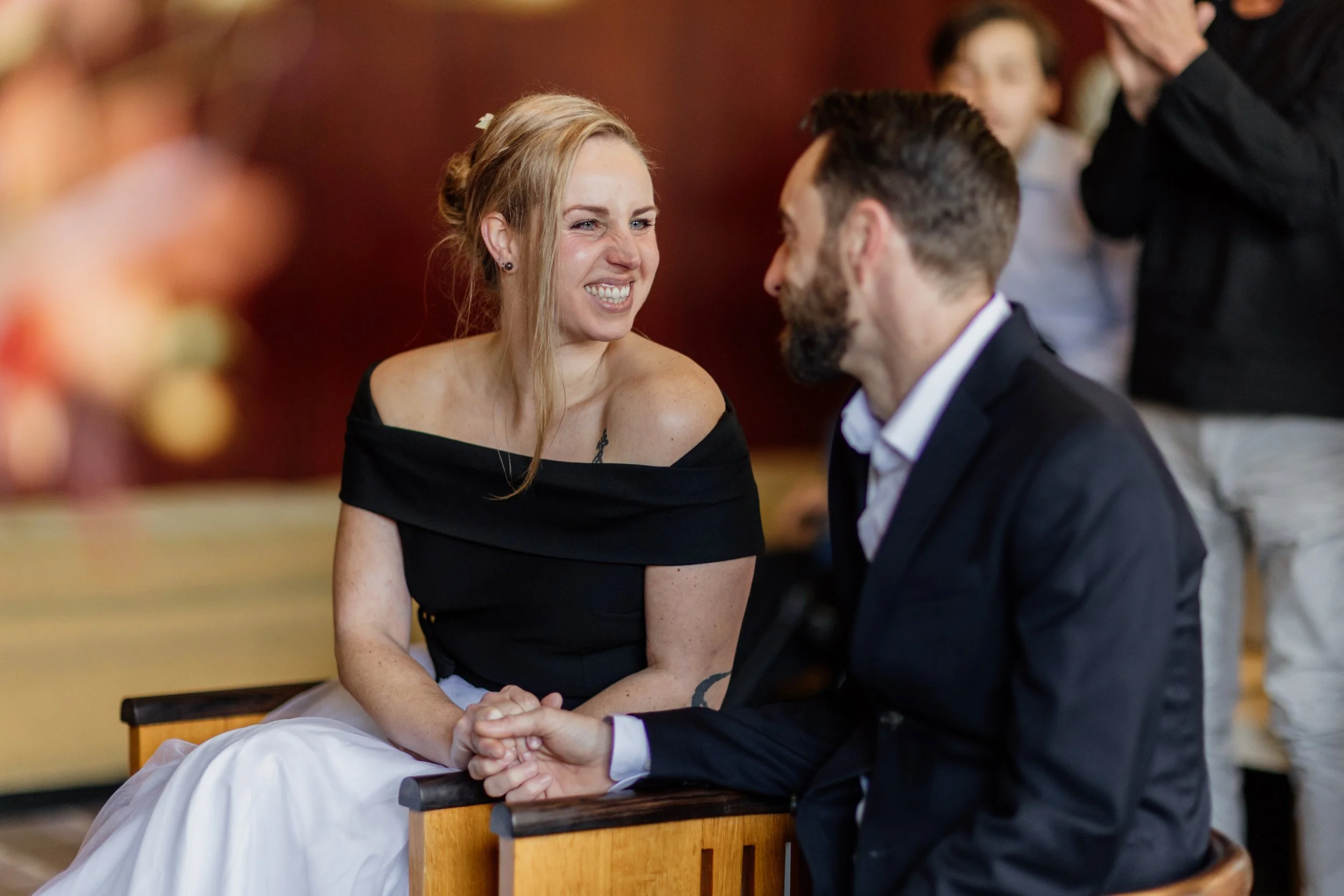 Een vrouw en een man zitten samen hartstochtelijk te knuffelen en te lachen tijdens een huwelijksceremonie, met anderen op de achtergrond.