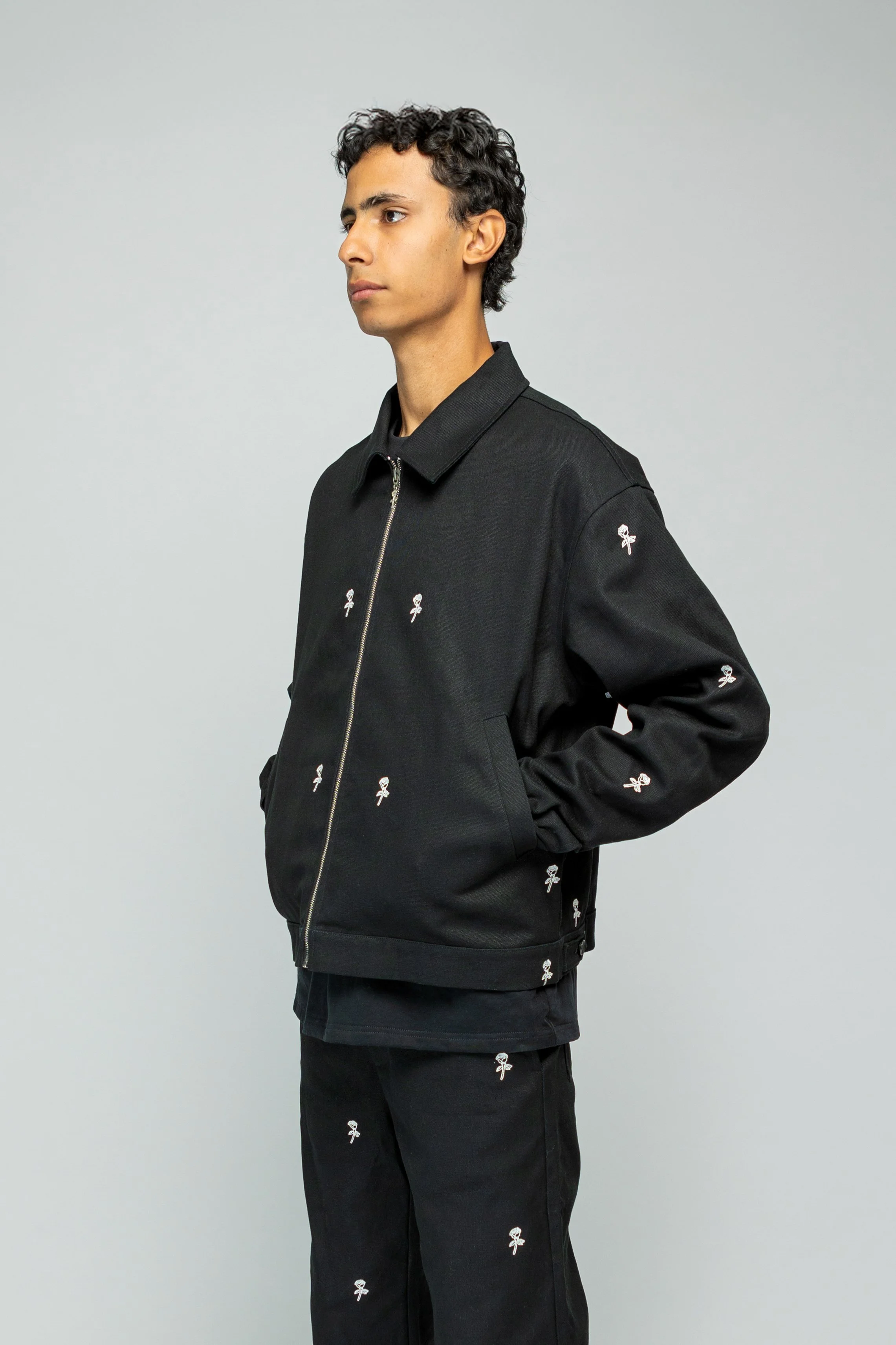 Man draagt een zwarte jas en broek, allebei versierd met kleine roze en witte bloemmotieven, staand tegen een lichte achtergrond.