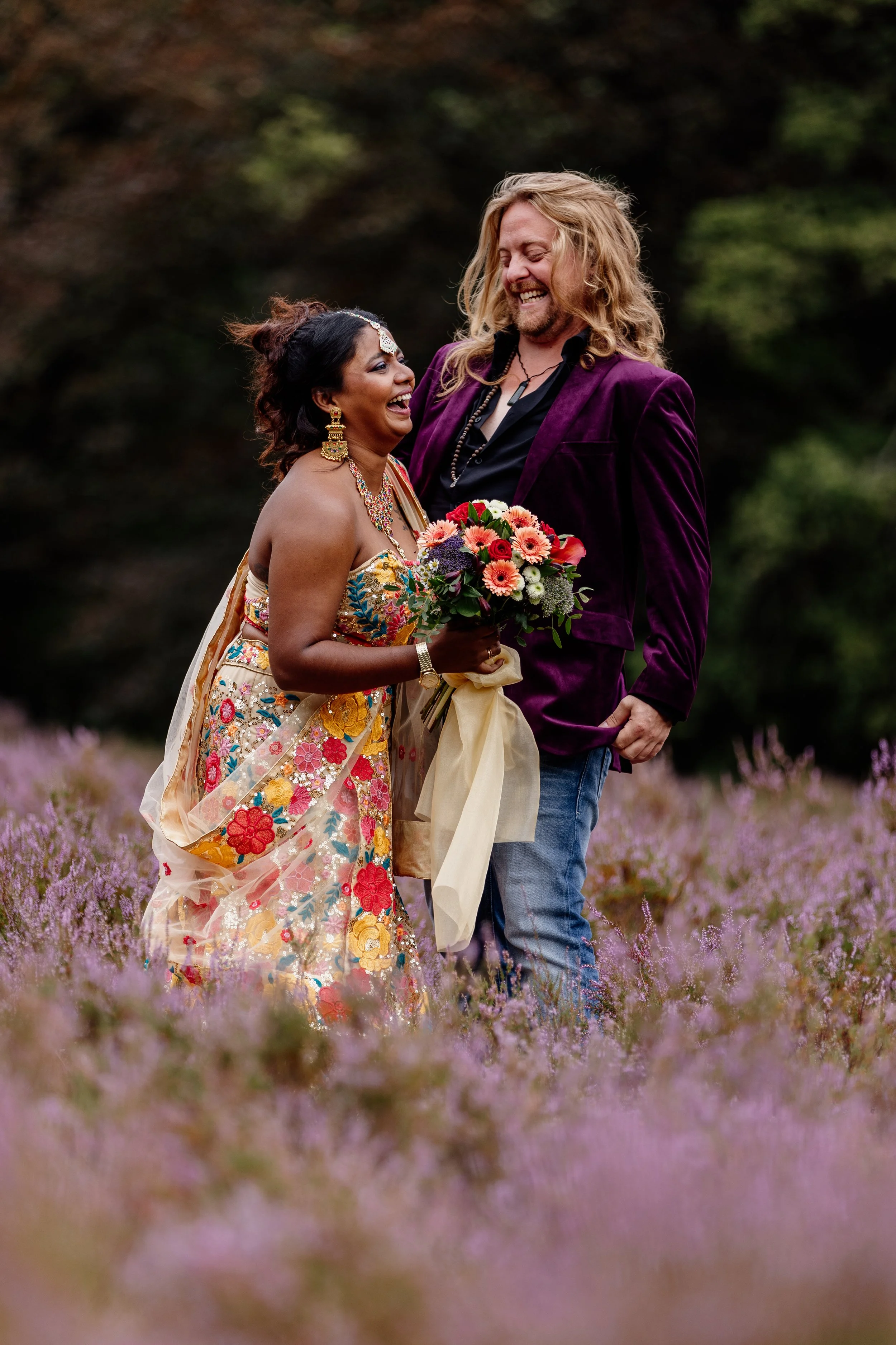 Twee mensen lachen en dansen in een veld vol paarse bloemen, een vrouw in een traditionele, kleurrijke jurk en een man met lang, golvend haar in een paarse jas.