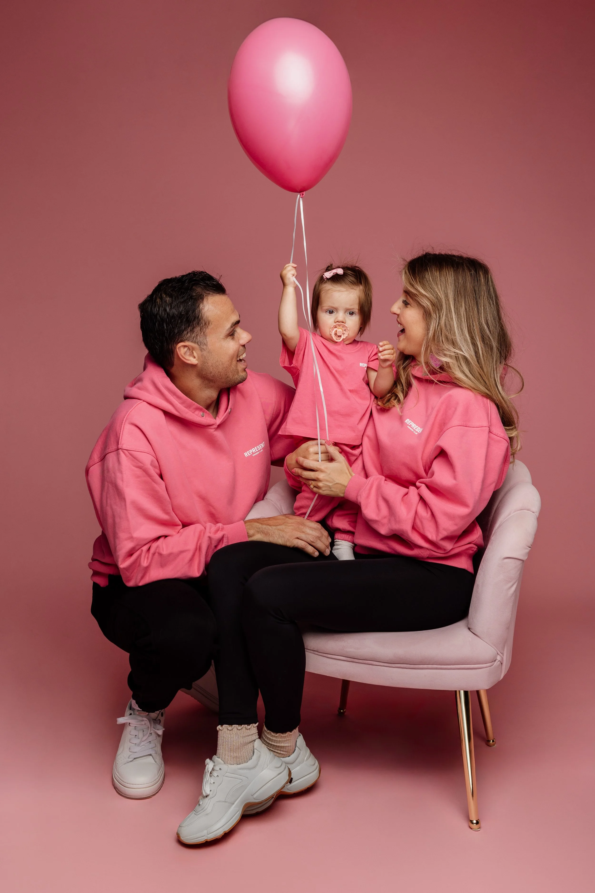 Gezin met twee volwassenen en een baby, allen gekleed in roze hoodie, zitten op een roze achtergrond. Ze lachen en spelen met een roze ballon.